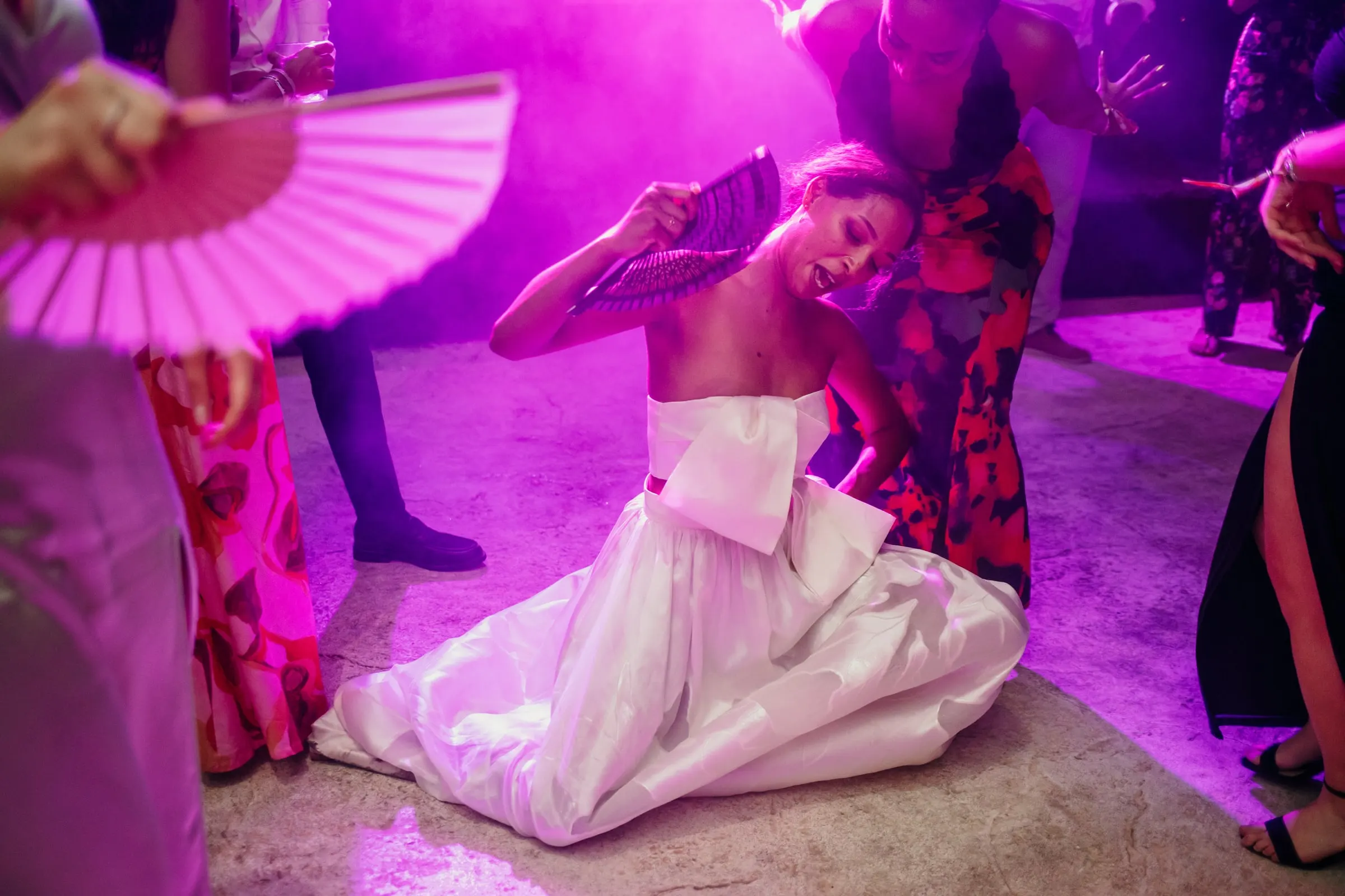 La novia baila en el suelo mientras una invitada agita un abanico rosa bajo luz magenta