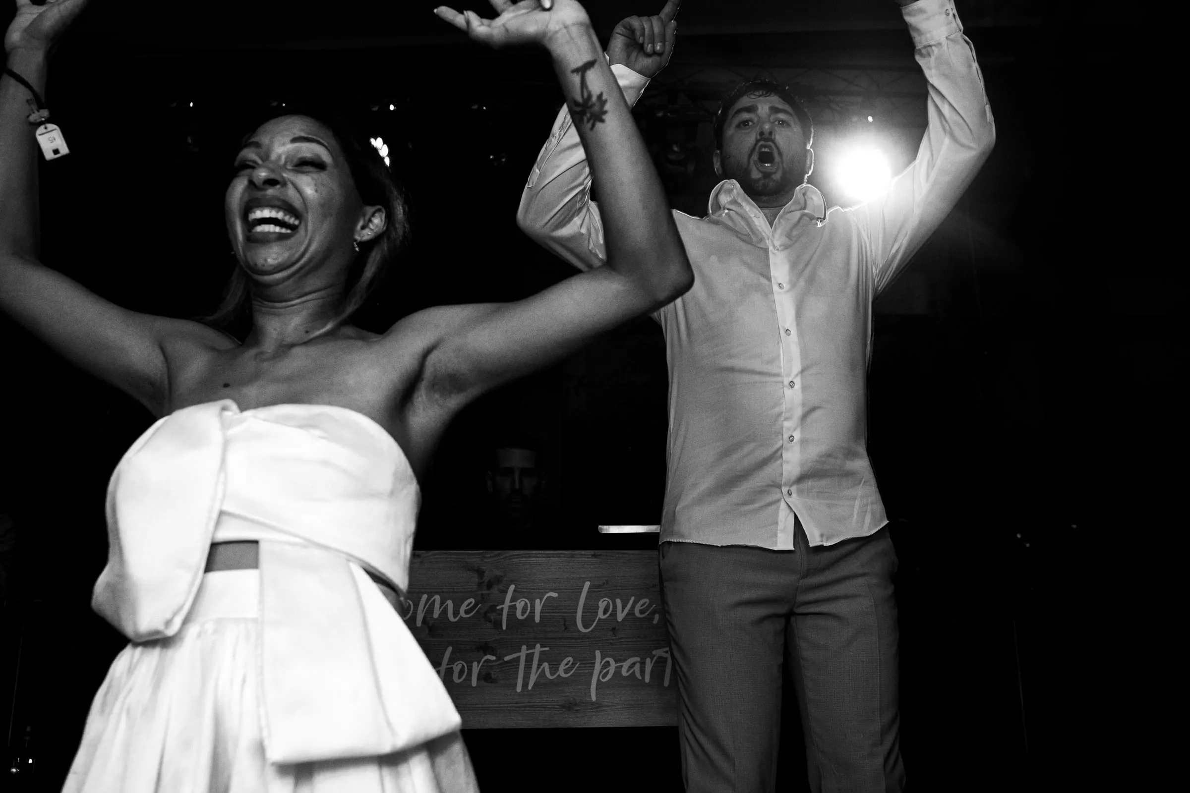 Foto en blanco y negro de los novios con los brazos en alto ante un cartel de 'come for love, stay for the party'