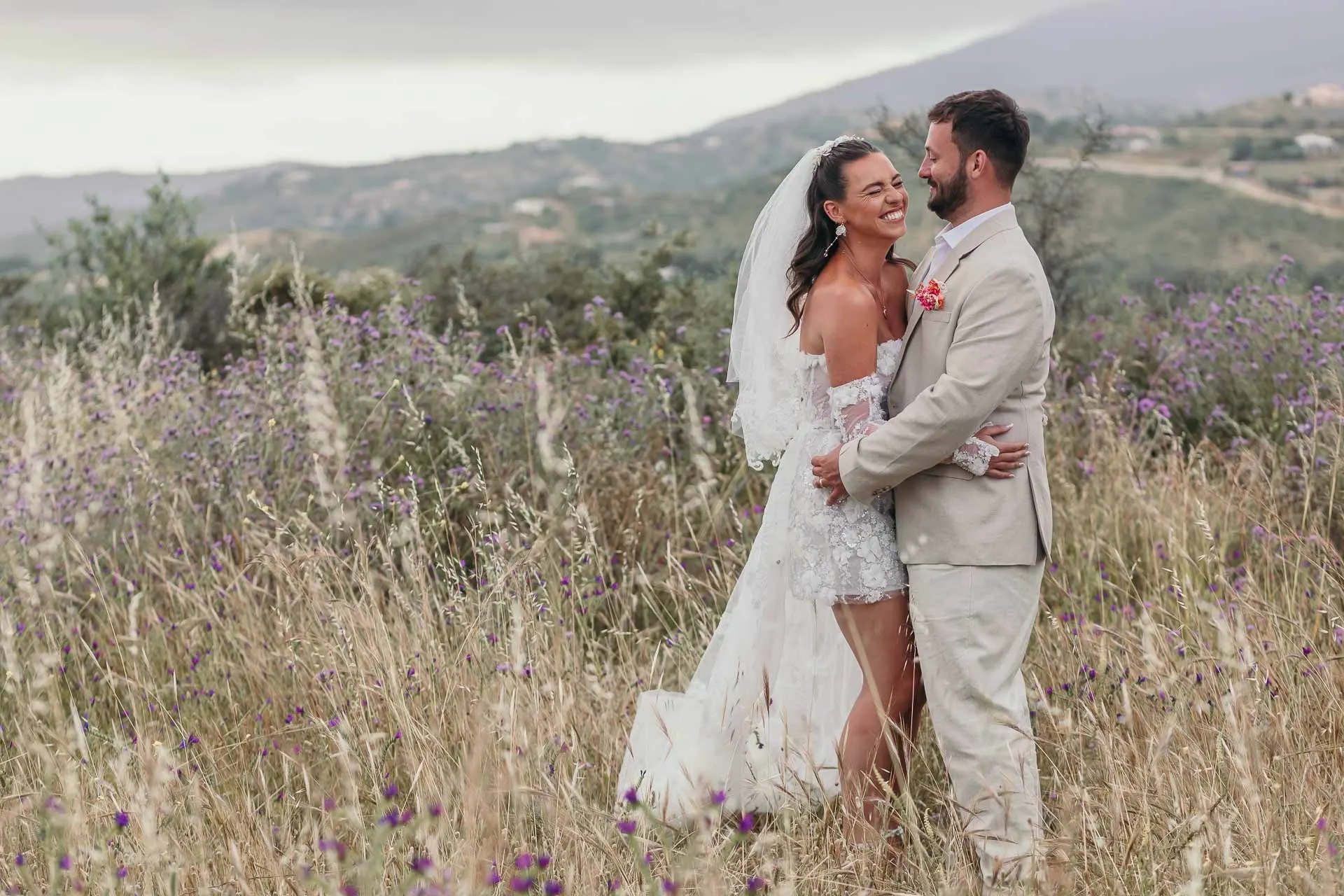 Los novios abrazados entre hierbas altas y flores moradas, con las colinas al fondo, Hacienda San José, Mijas