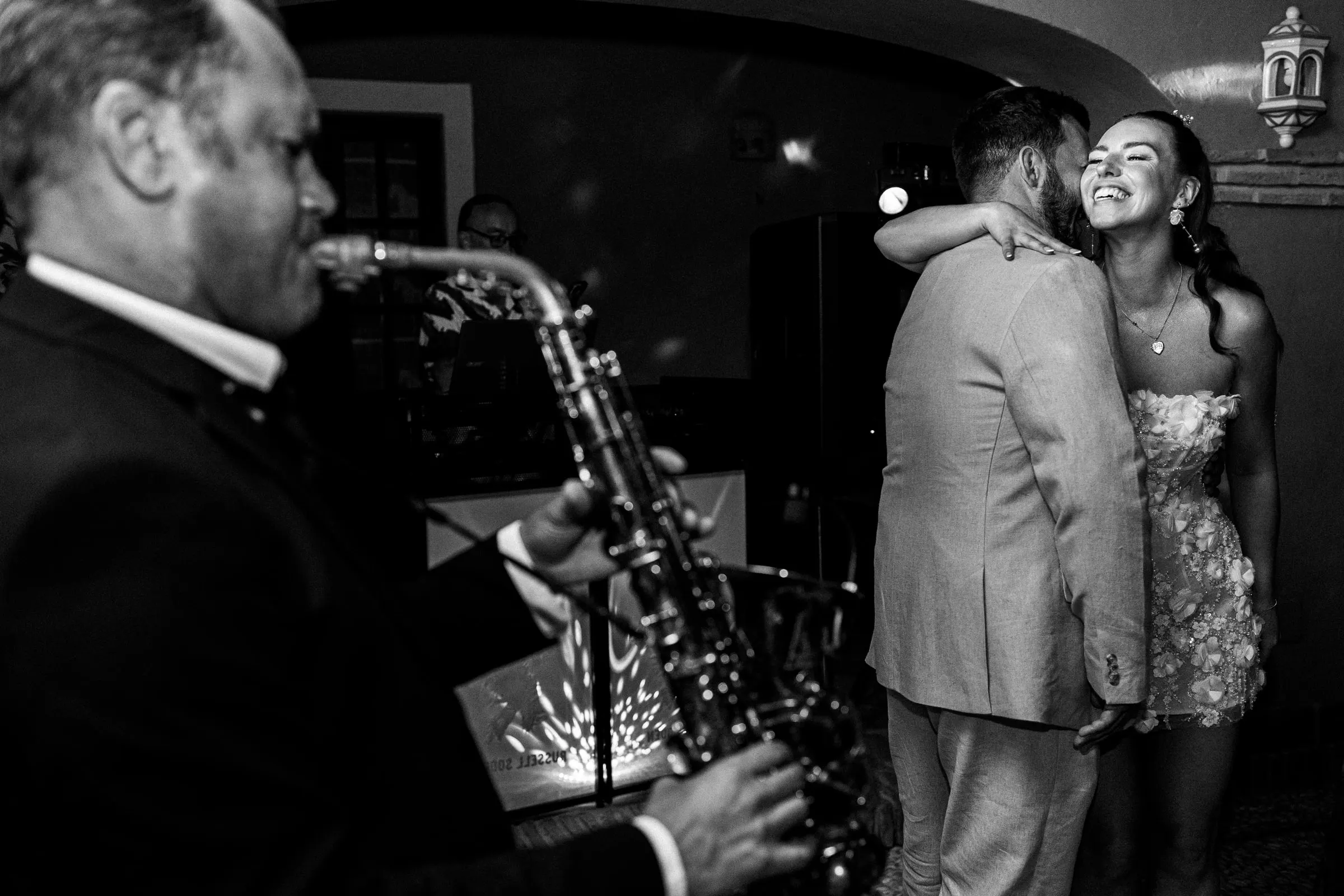La novia abraza al novio en la pista mientras toca un saxofonista, en blanco y negro, Hacienda San José, Mijas