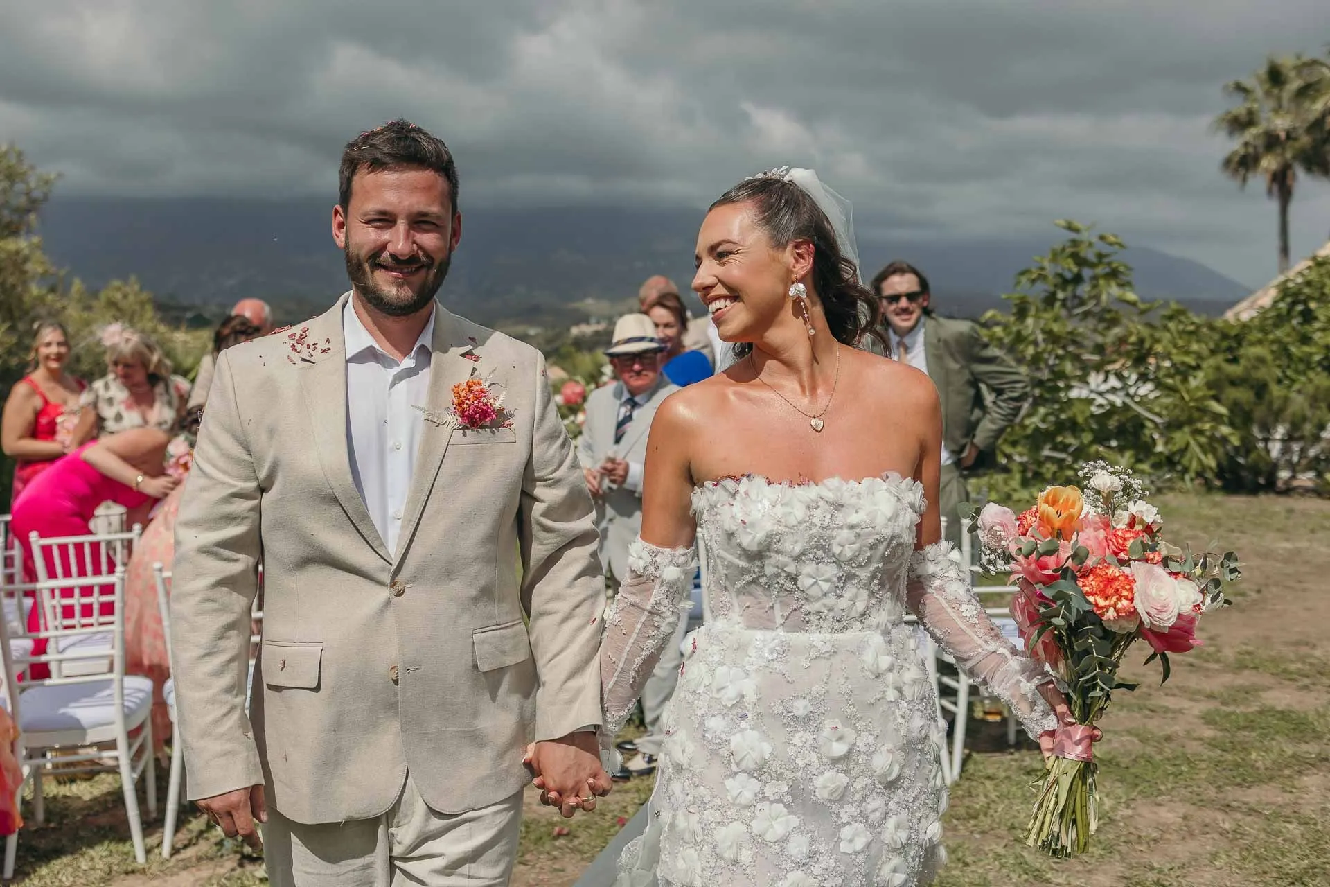 Los novios salen de la mano por el pasillo, sonrientes, con los invitados de rosa detrás, Hacienda San José, Mijas