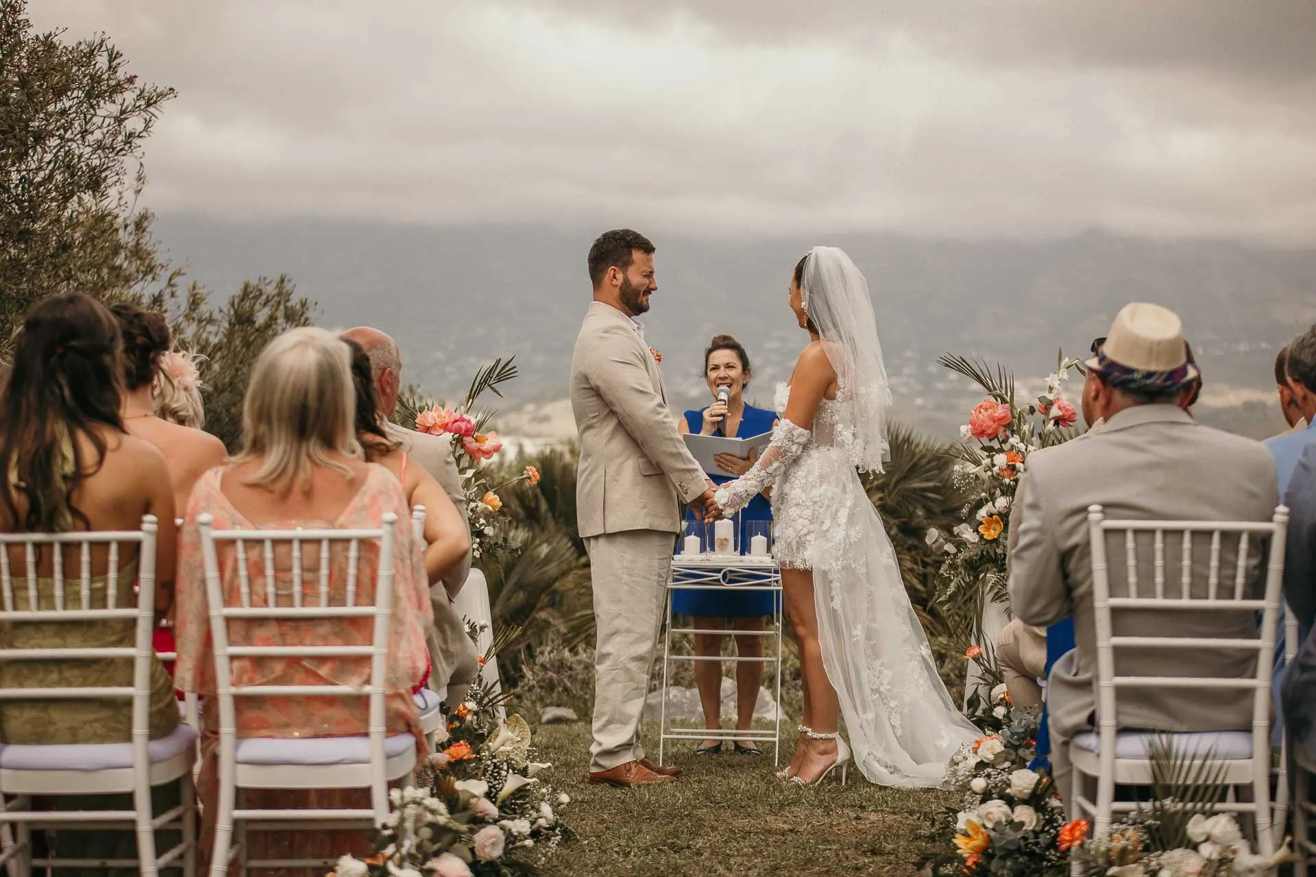 Bride and groom hold hands before the celebrant at the hilltop ceremony, guests seated, Hacienda San José, Mijas