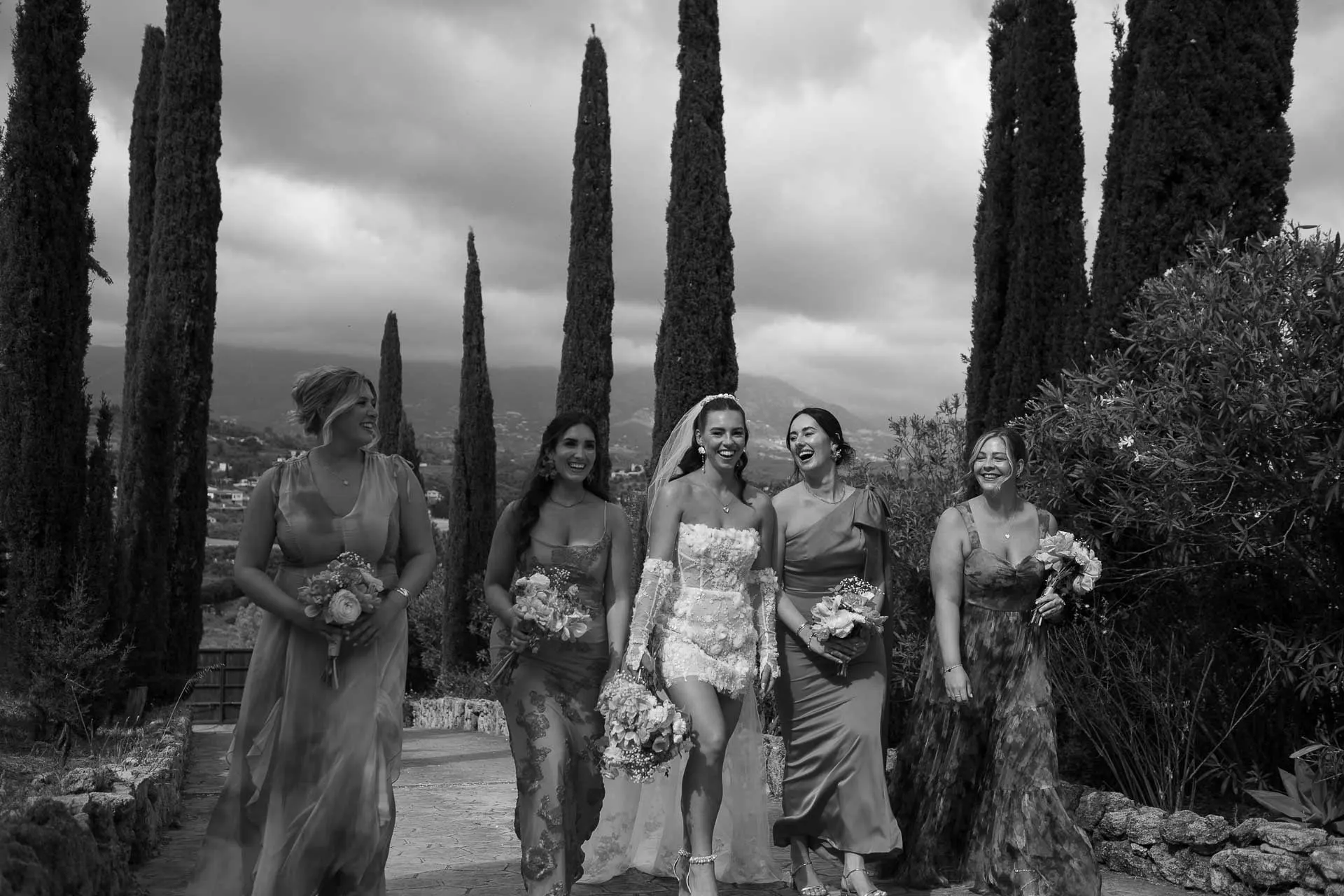 La novia camina con sus damas de honor entre cipreses altos, en blanco y negro, Hacienda San José, Mijas