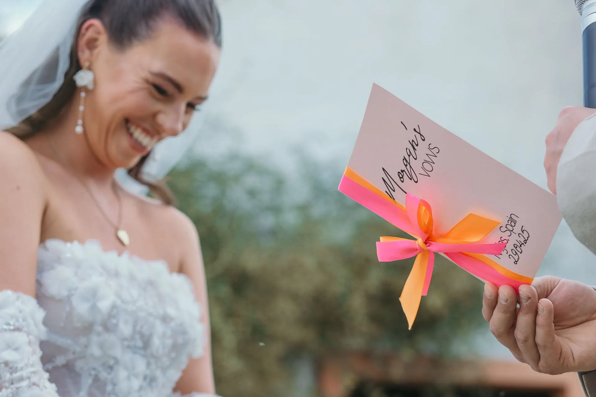 La novia se ríe al ver la tarjeta de votos del novio atada con lazo rosa, Hacienda San José, Mijas