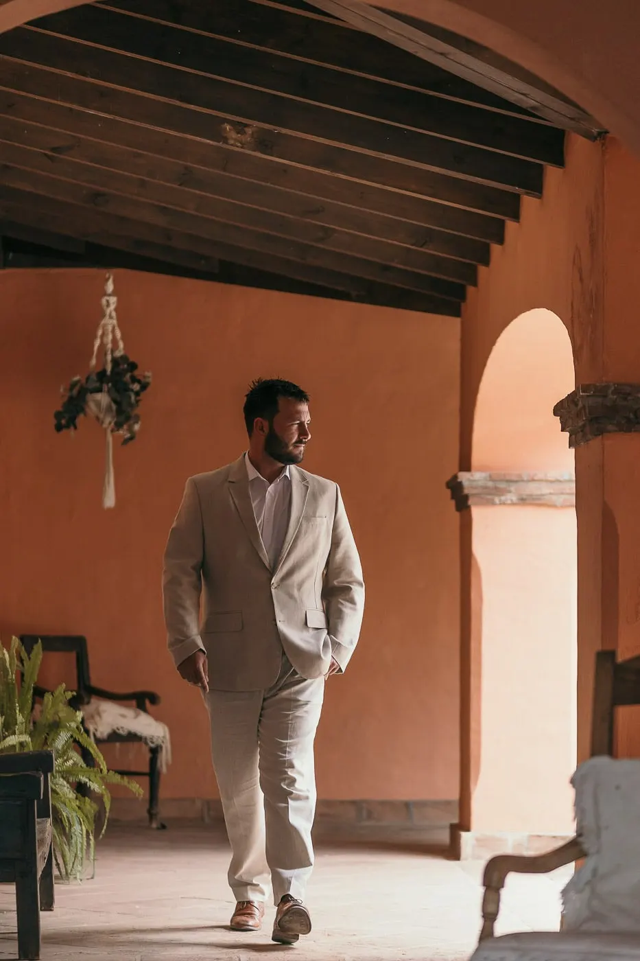 Groom in a beige suit walking through an arched terracotta porch, hands in pockets, Hacienda San José, Mijas