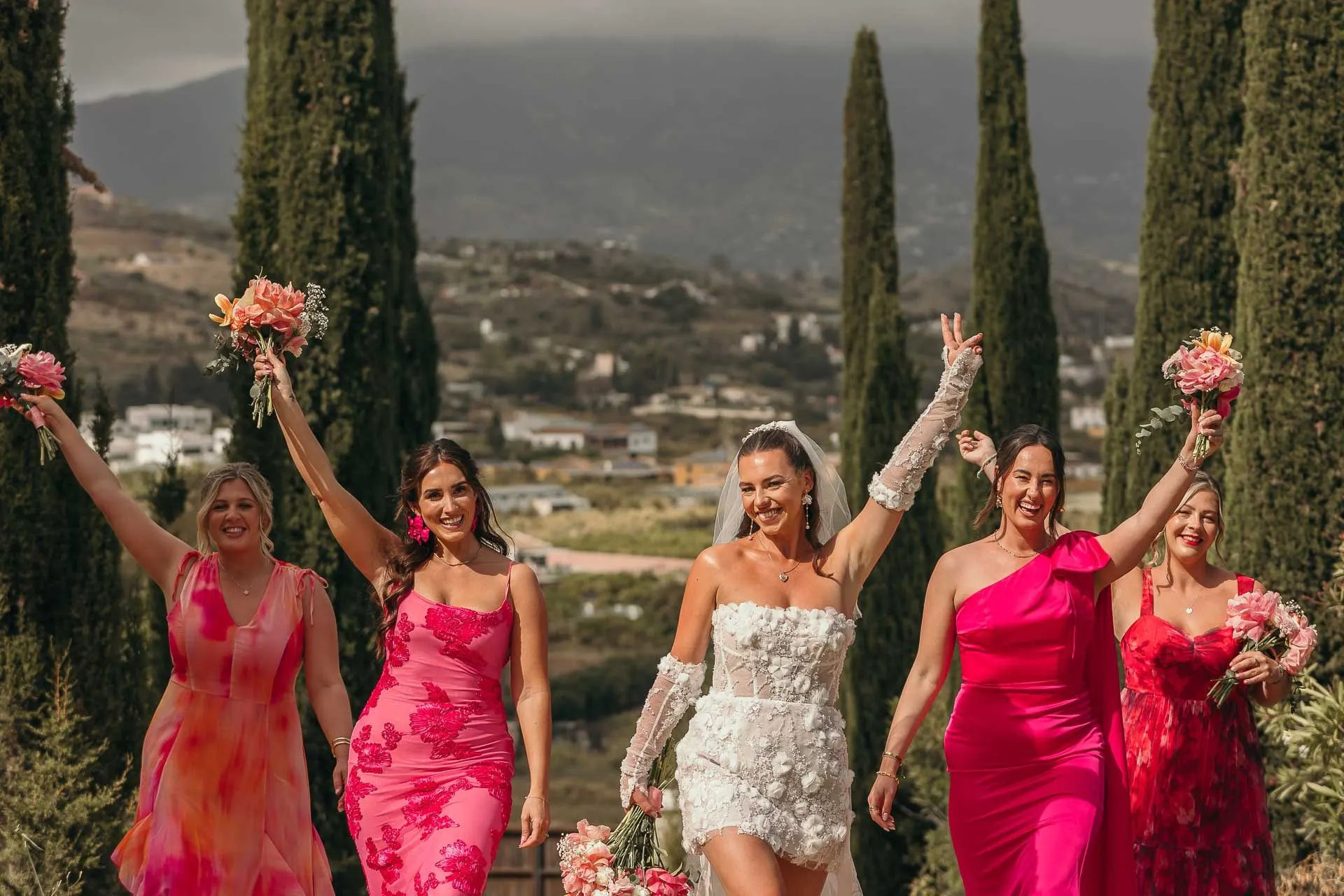 Bride and bridesmaids in pink cheering with bouquets raised between tall cypresses, Hacienda San José, Mijas