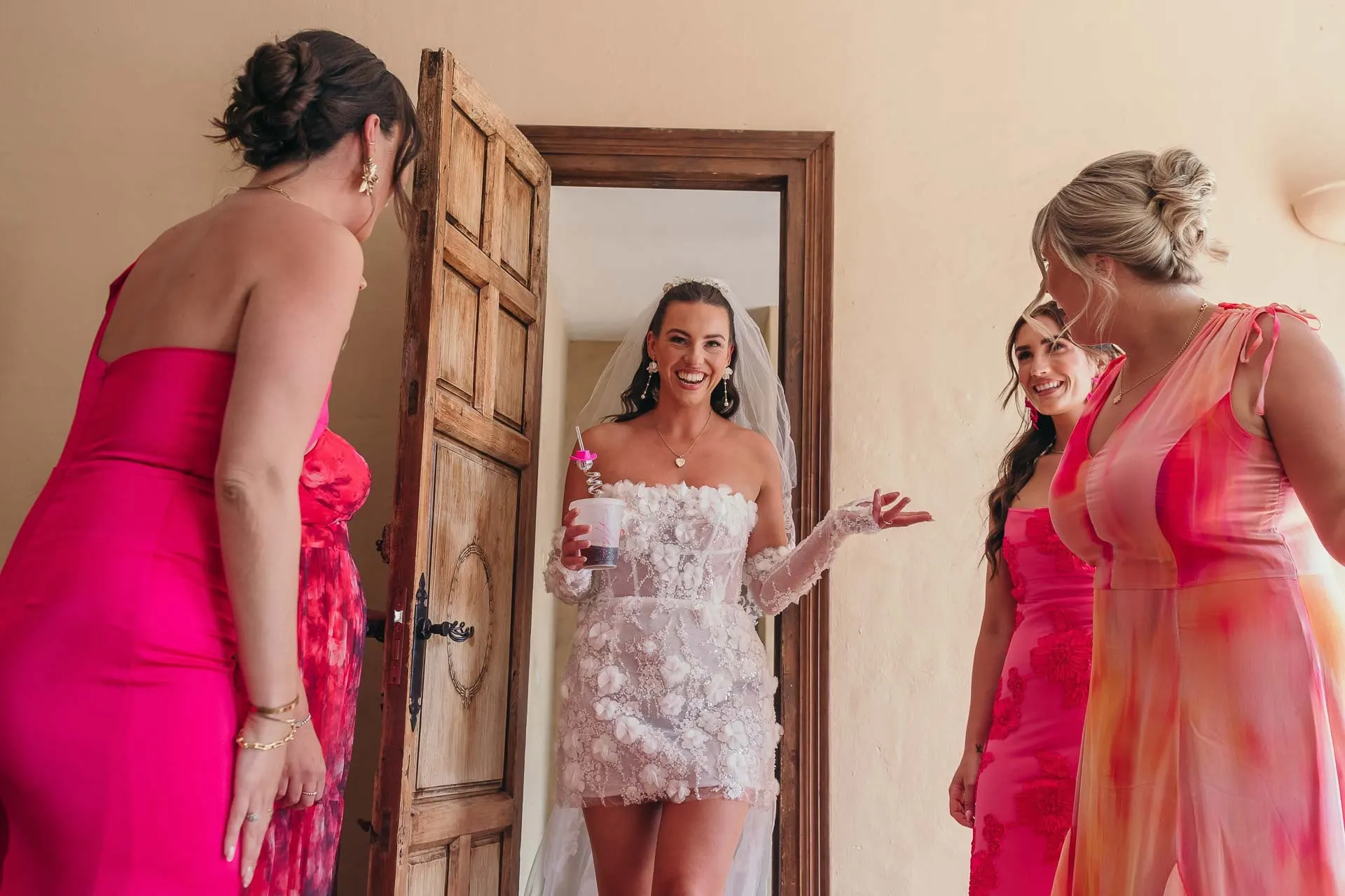 Bride steps through a wooden door to meet her bridesmaids in pink, all smiling, Hacienda San José, Mijas