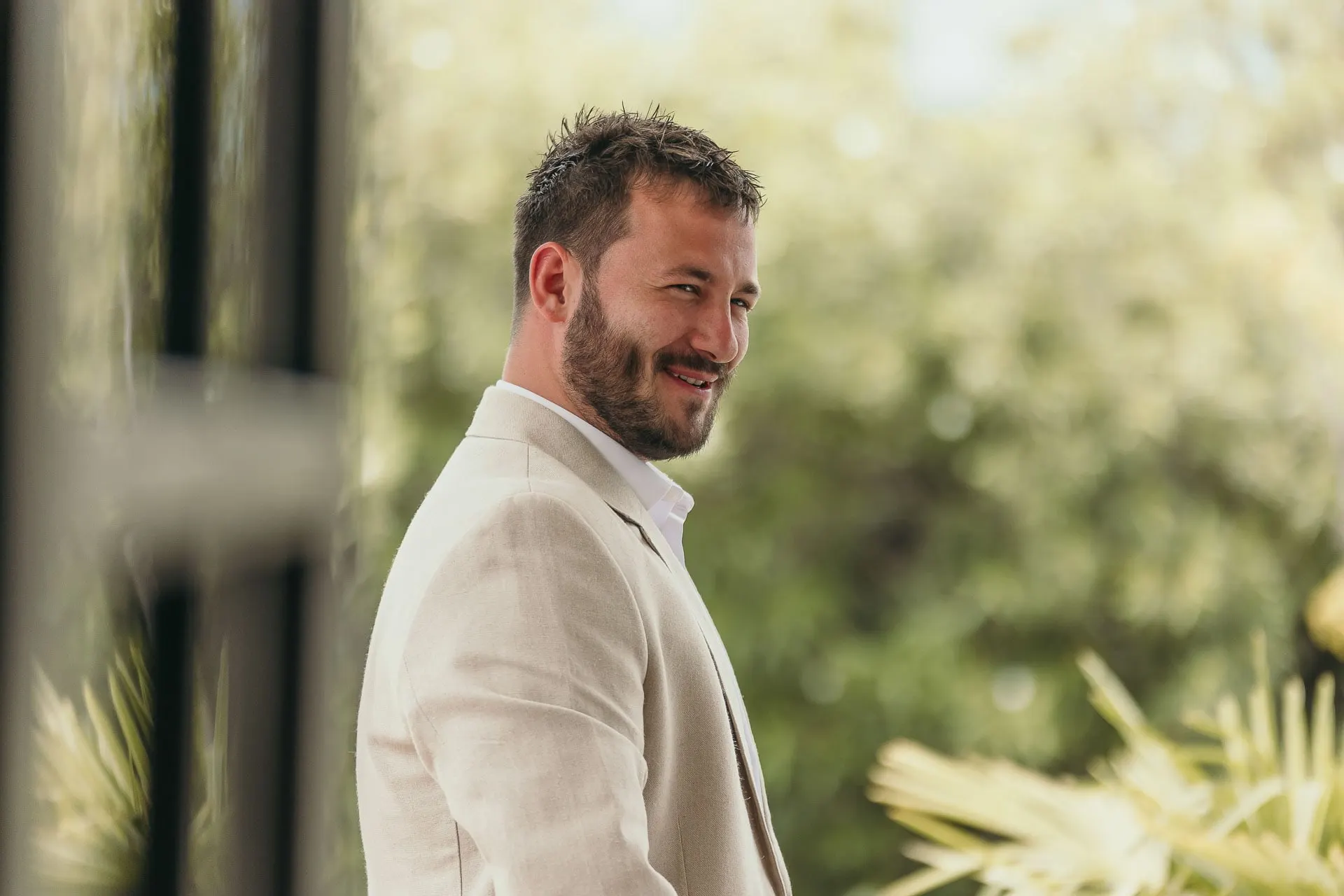 Groom in a beige linen jacket smiling to one side in the garden, Hacienda San José, Mijas