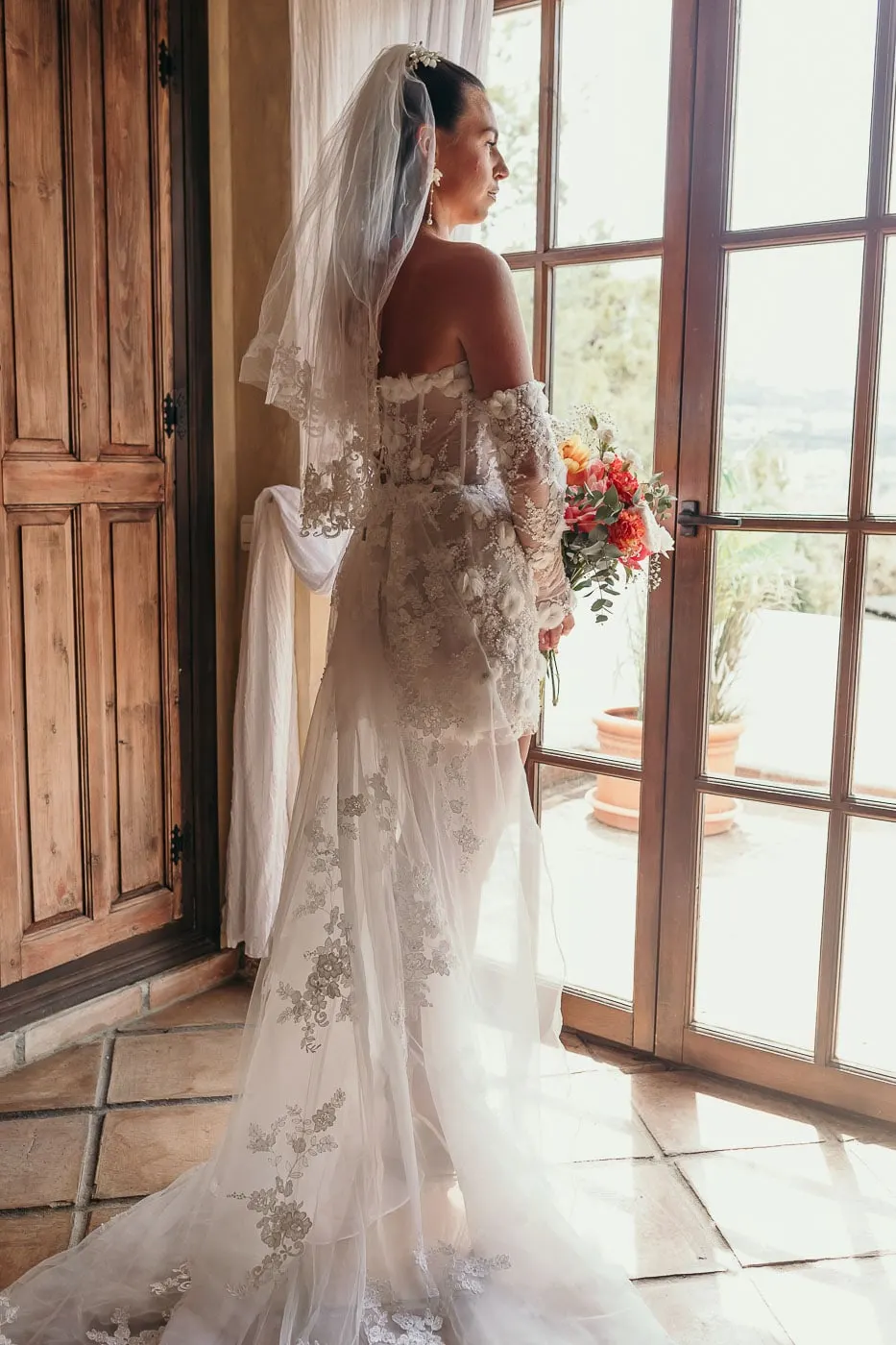 Bride with veil and bouquet looking out of tall glazed doors, her lace train on the tiles, Hacienda San José, Mijas