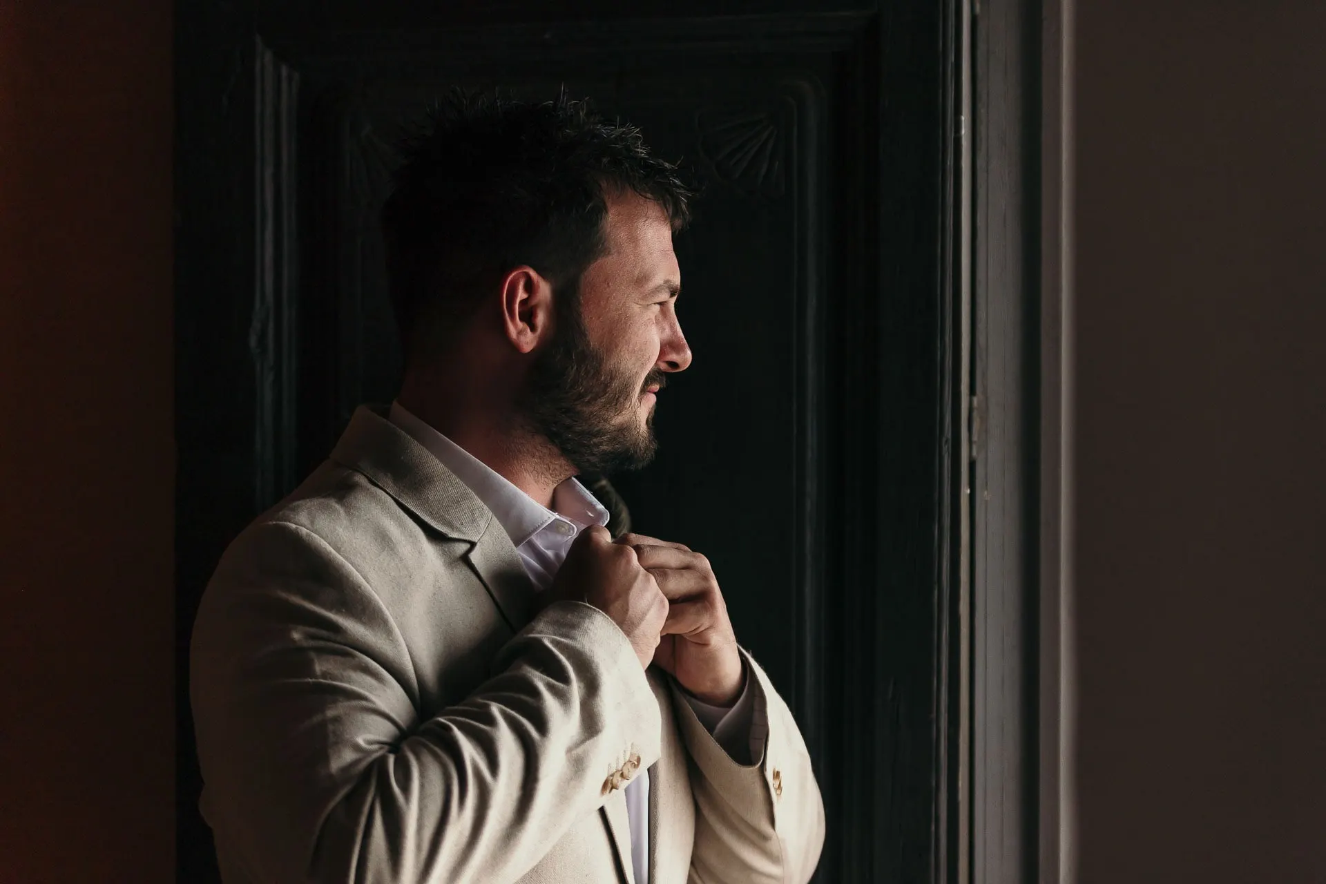 Groom buttoning his beige jacket in a dark doorway, looking out to the light, Hacienda San José, Mijas