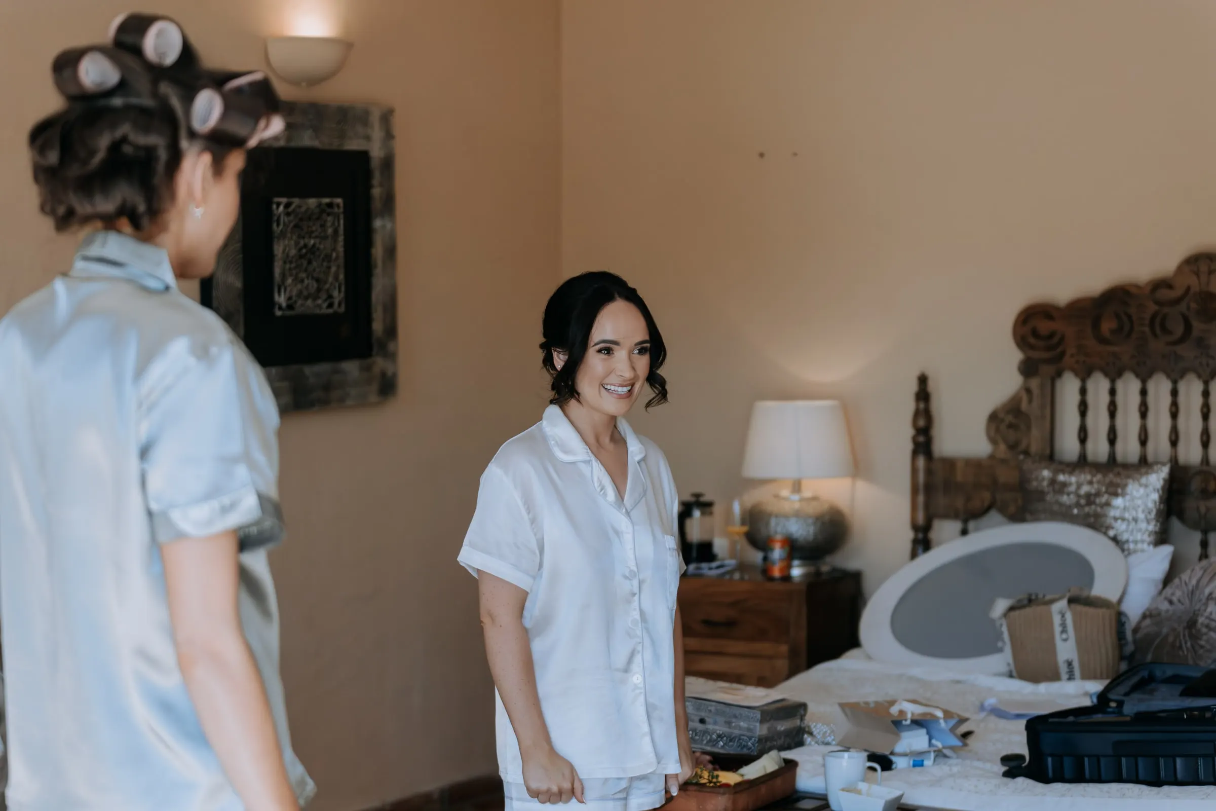 Bride and a bridesmaid in rollers chatting in the bedroom with the carved wooden headboard, Mijas