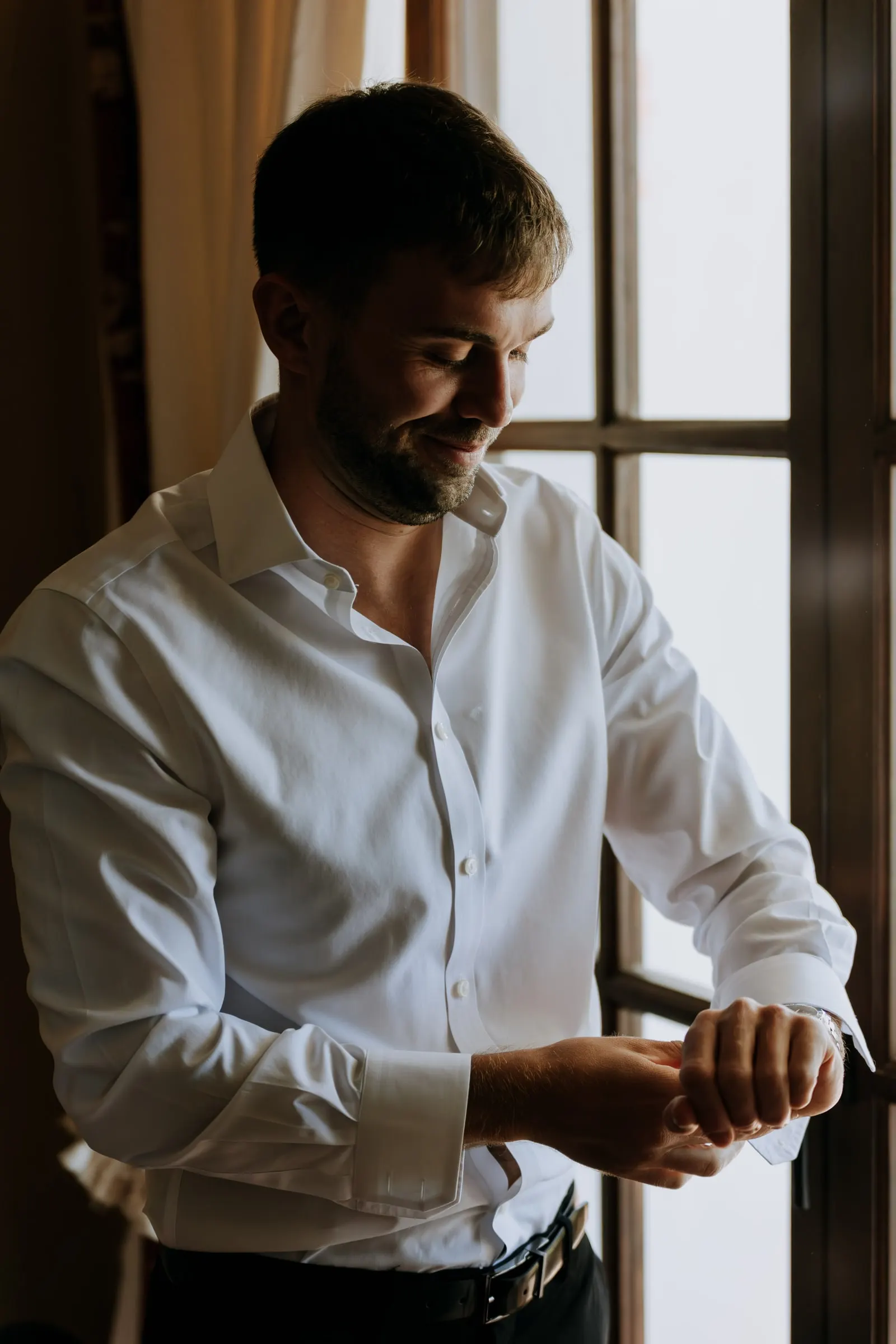 Groom fastening his cuffs by a tall window, Mijas