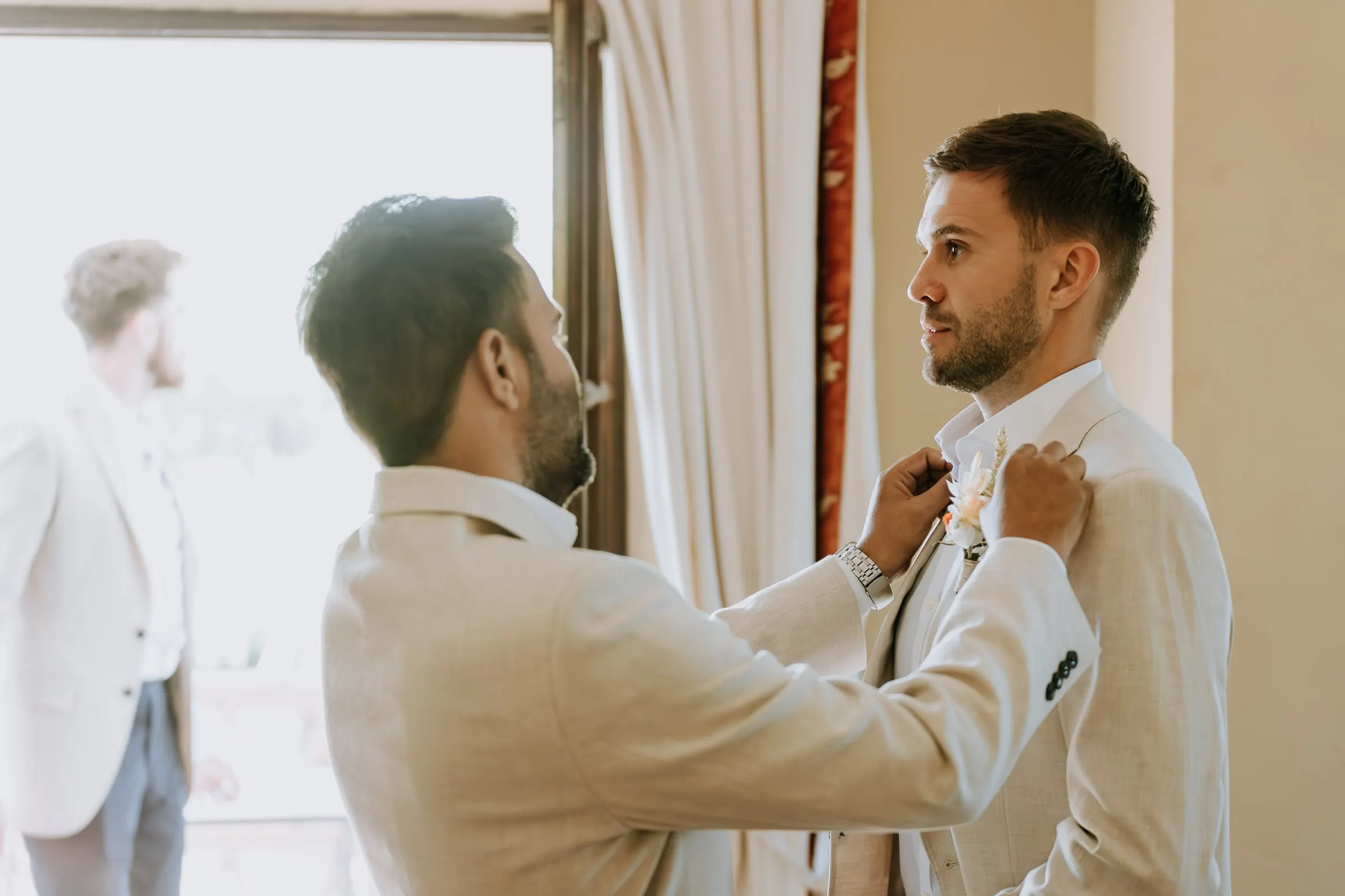 Friend pinning a dried-flower buttonhole on the groom by the window, Hacienda San José
