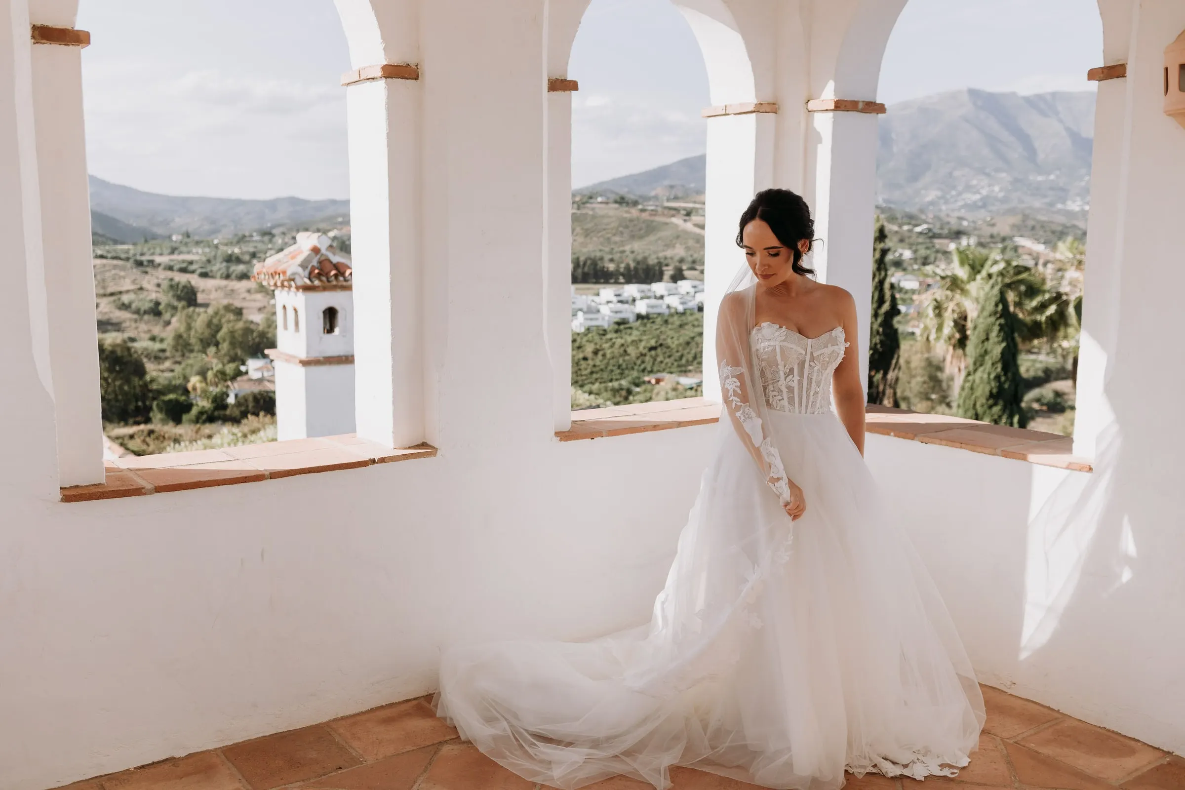 Bride standing among the white arches of the tower, with the bell tower and the Sierra de Mijas behind