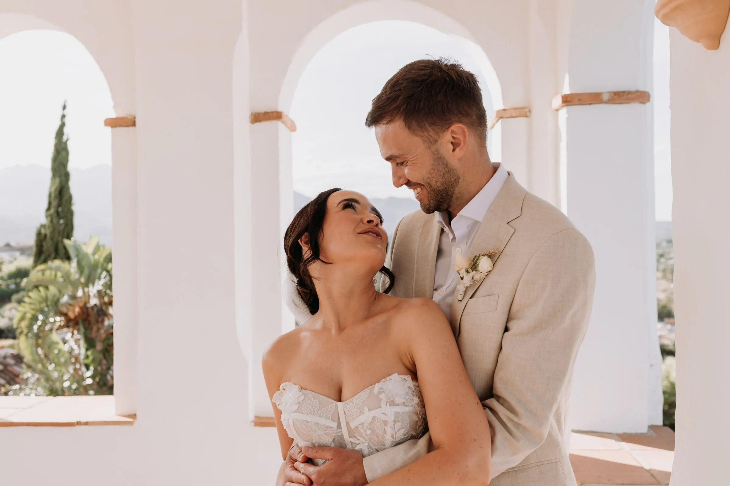 Groom hugging the bride from behind under the white arches, Hacienda San José