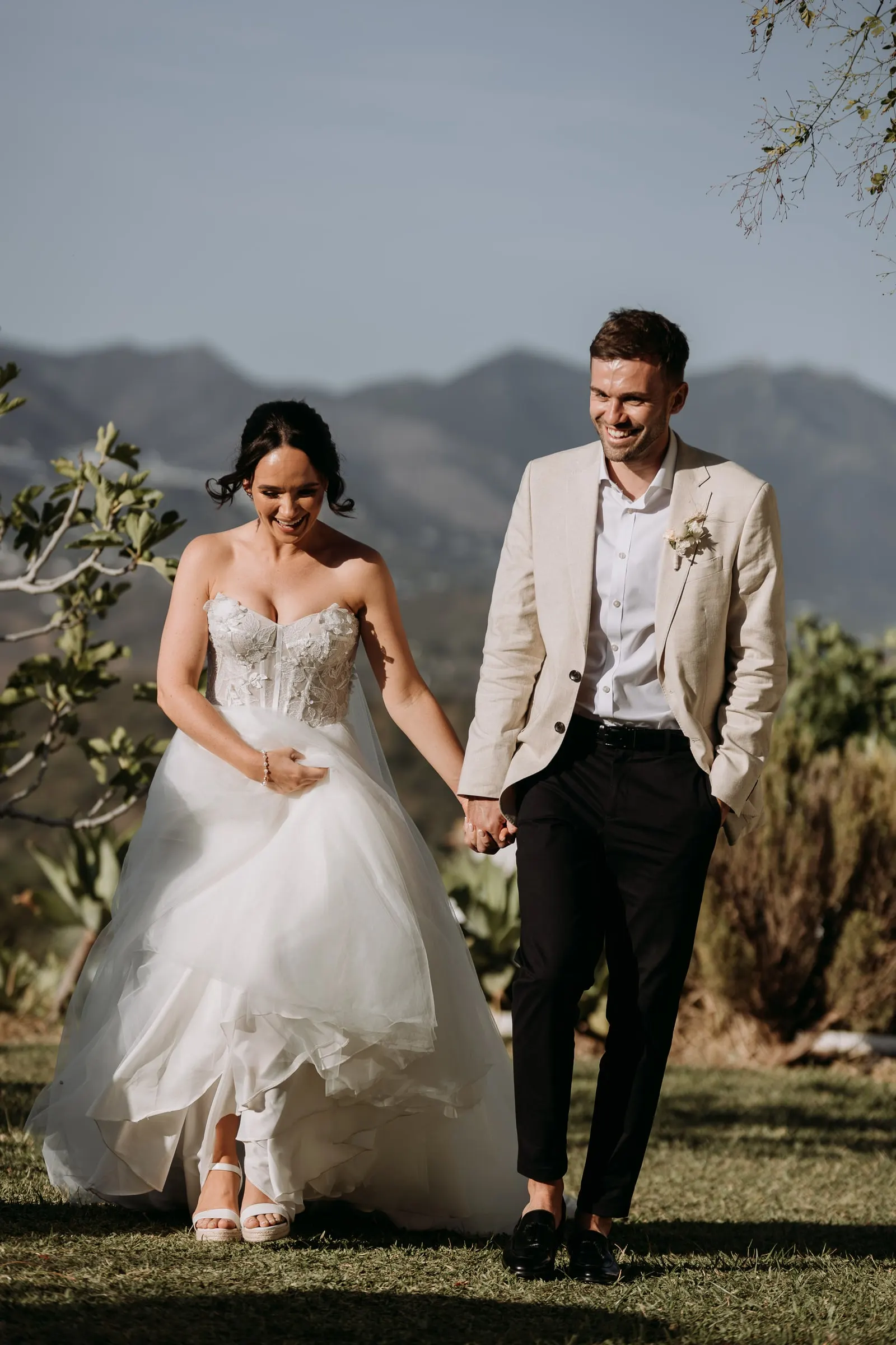Bride and groom walking hand in hand across the lawn with the Sierra de Mijas behind