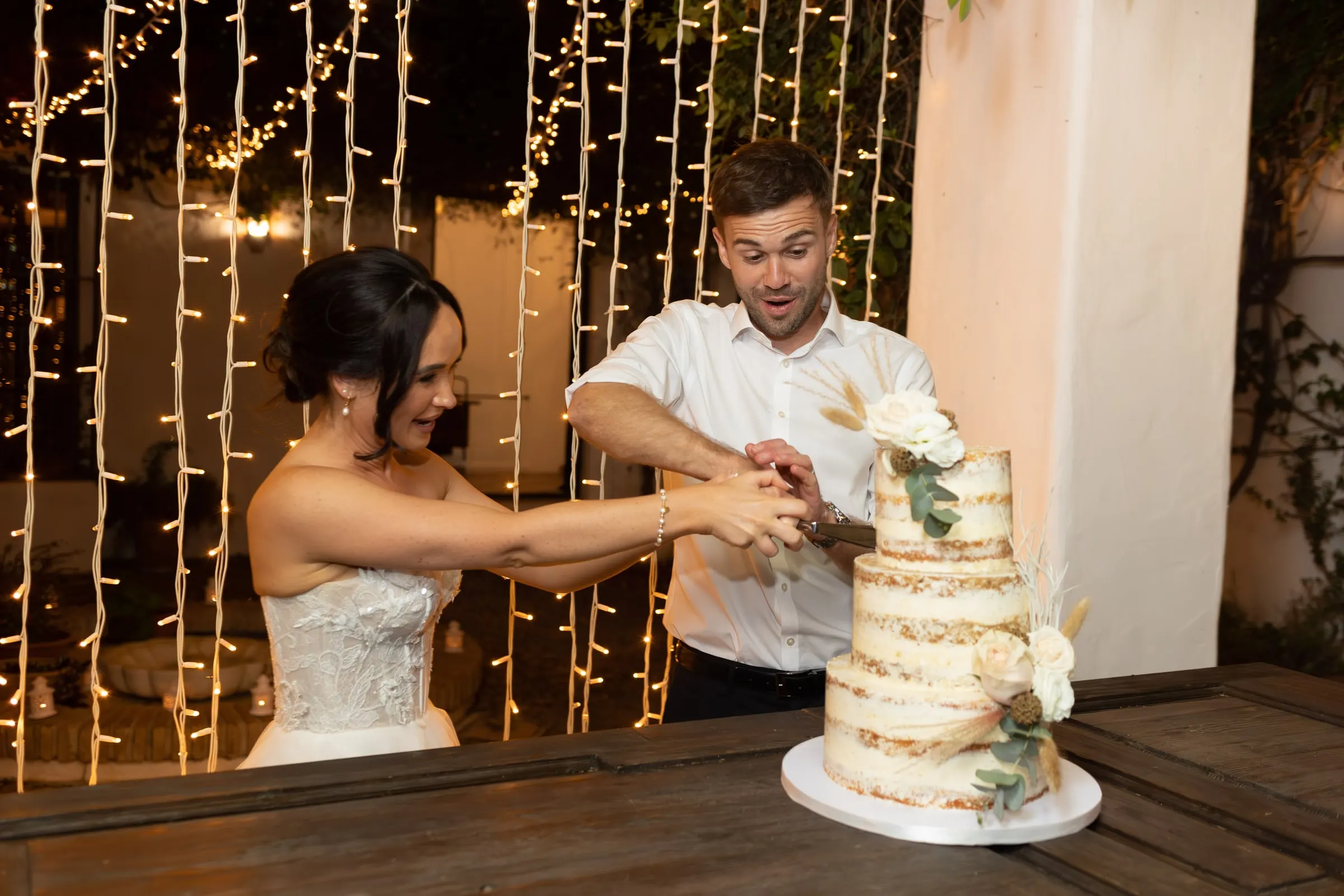 Bride and groom cutting a naked cake under a curtain of lights, Mijas