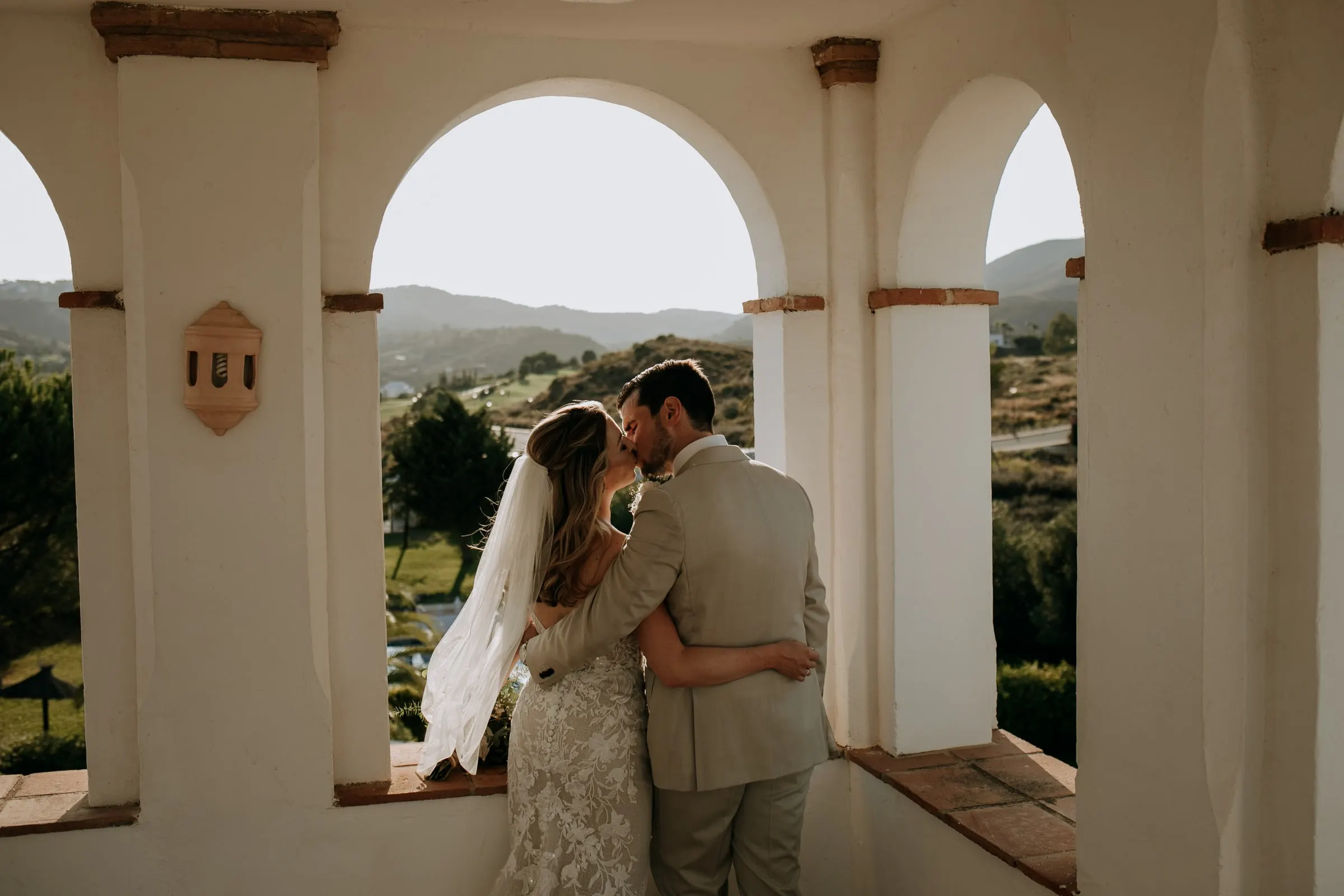 Los novios se besan bajo un arco blanco de la galería, con las colinas al fondo, Hacienda San José, Mijas