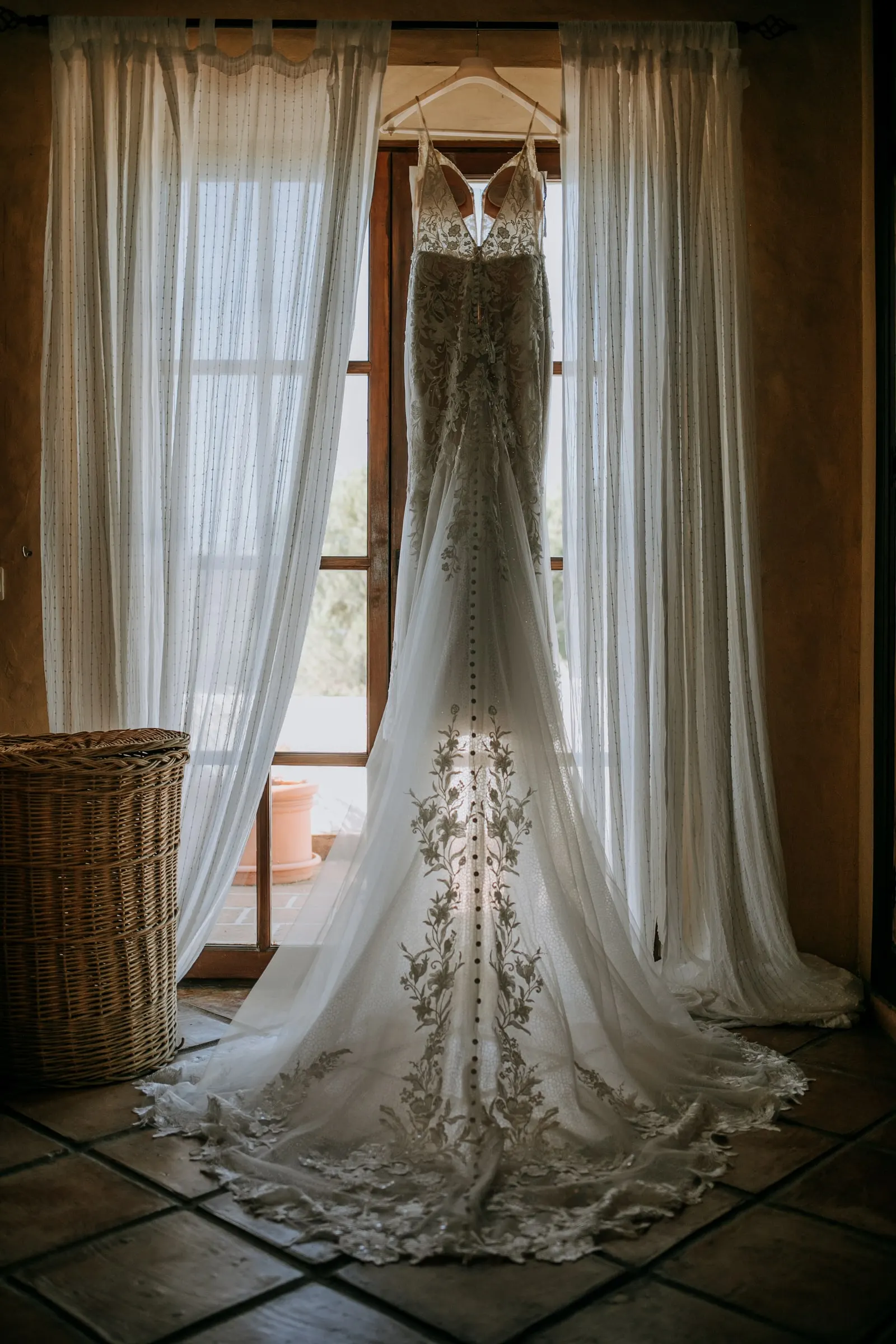 Vestido de novia de encaje con cola larga colgado ante una ventana con visillos, Hacienda San José, Mijas