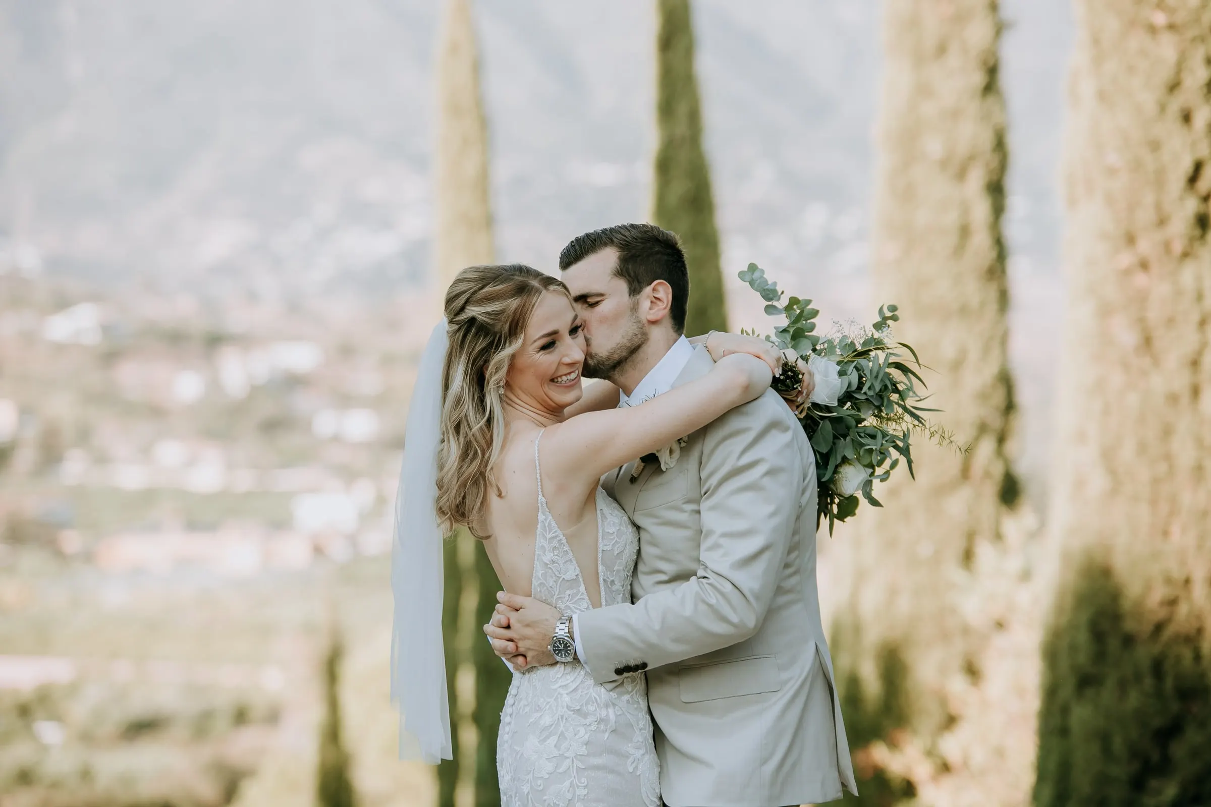 El novio besa la mejilla de la novia, que sonríe, con cipreses y el valle detrás, Hacienda San José, Mijas