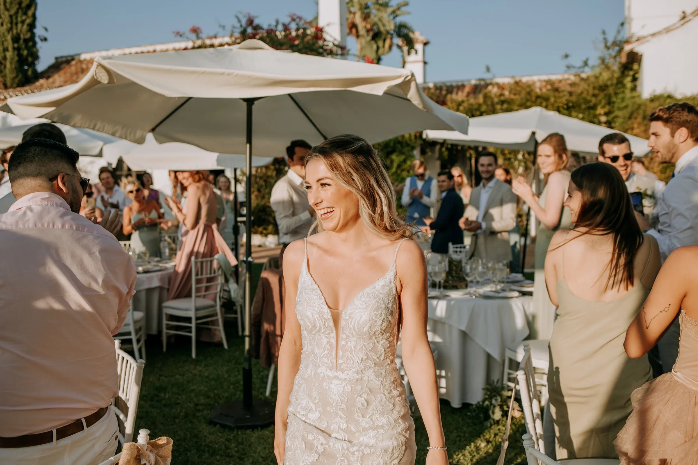 La novia sonríe entre los invitados bajo sombrillas blancas en el cóctel del jardín, Hacienda San José, Mijas