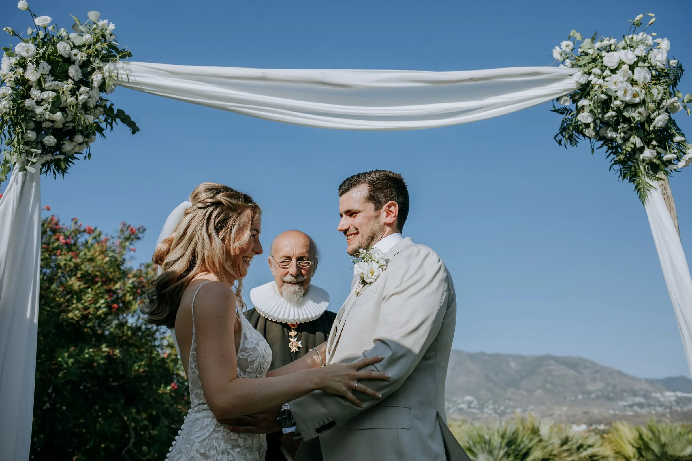 Los novios, de la mano ante el oficiante bajo un arco con flores y las montañas detrás, Hacienda San José, Mijas