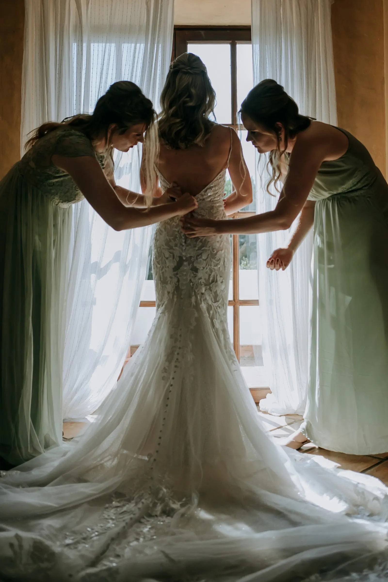 Dos damas de verde abrochan la espalda del vestido de la novia frente a una ventana luminosa, Hacienda San José, Mijas