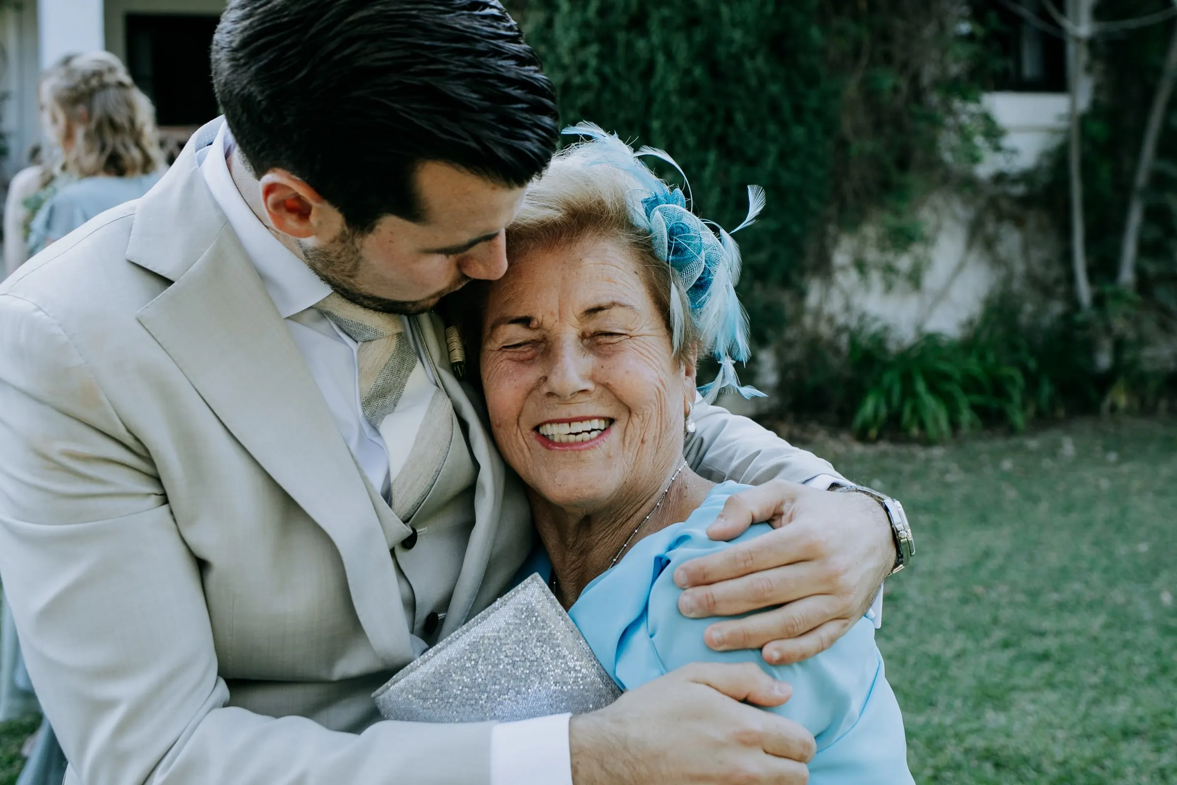 El novio abraza a una invitada mayor con tocado azul, que sonríe, en el césped, Hacienda San José, Mijas