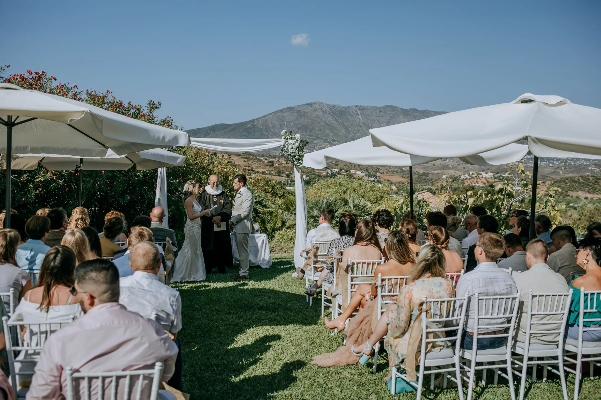 Invitados sentados en el césped bajo sombrillas blancas frente a la ceremonia, con la sierra detrás, Hacienda San José, Mijas
