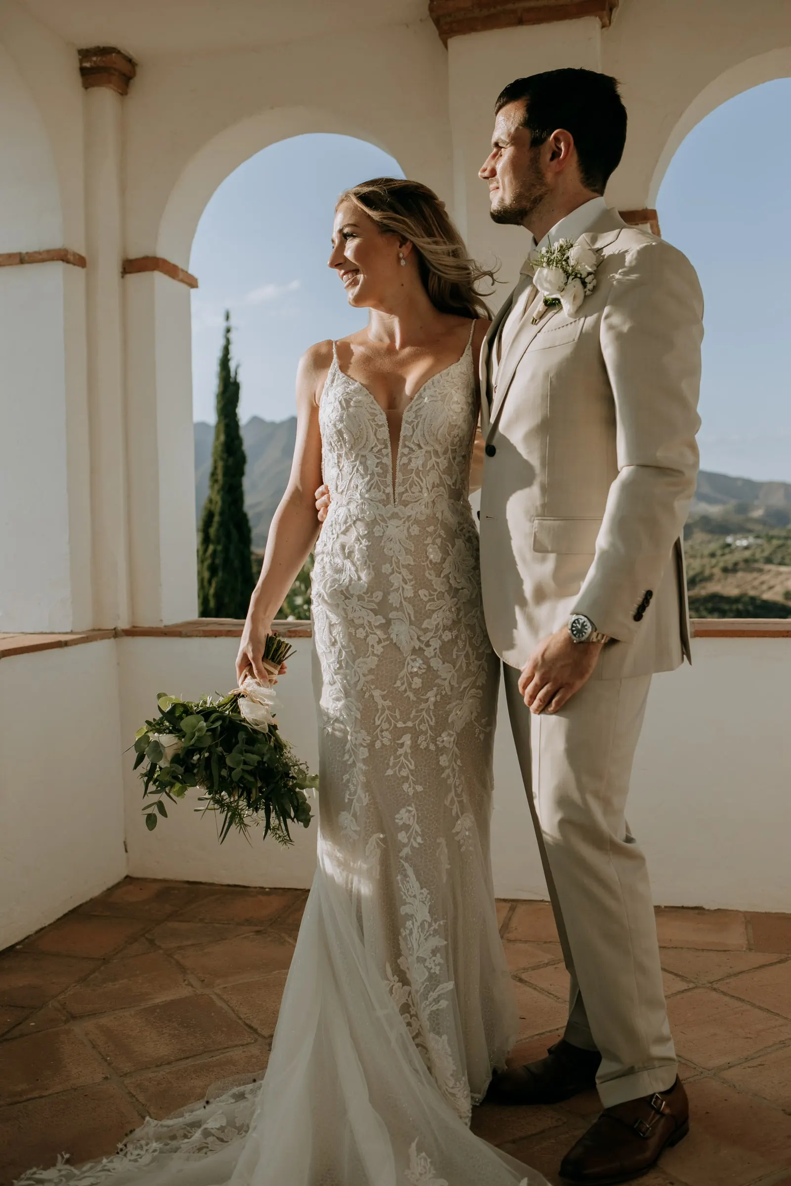 Los novios sonríen en la galería de arcos, ella con el ramo bajo, y las colinas detrás, Hacienda San José, Mijas