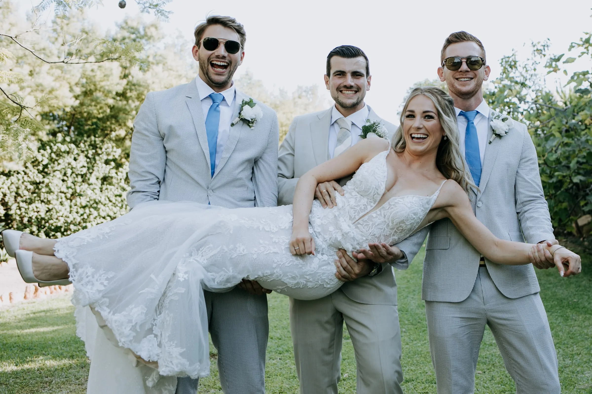 Tres amigos del novio con traje gris y gafas de sol levantan en brazos a la novia, que ríe, Hacienda San José, Mijas