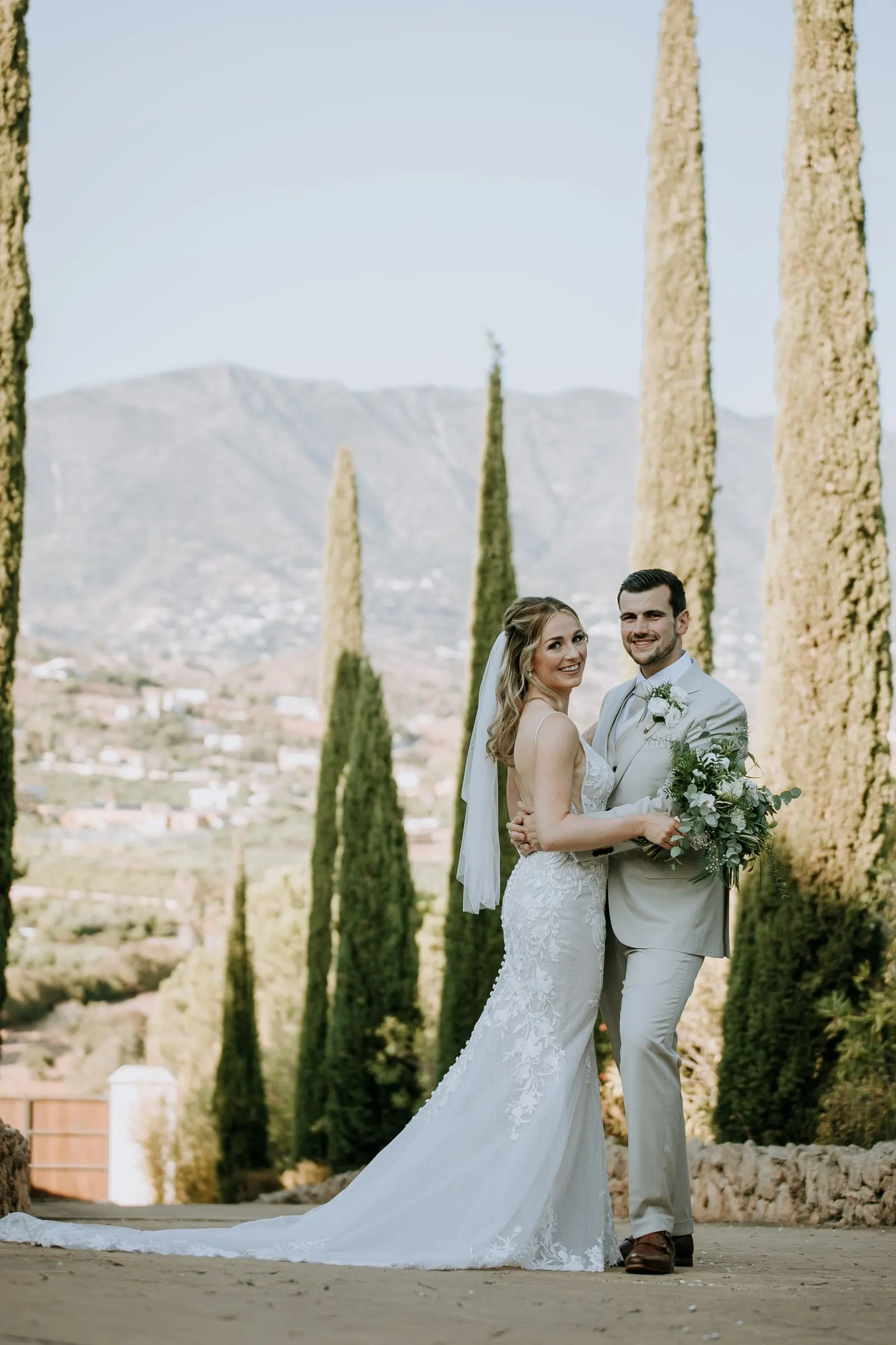 Los novios posan entre cipreses altos con la sierra detrás y la cola sobre el camino, Hacienda San José, Mijas