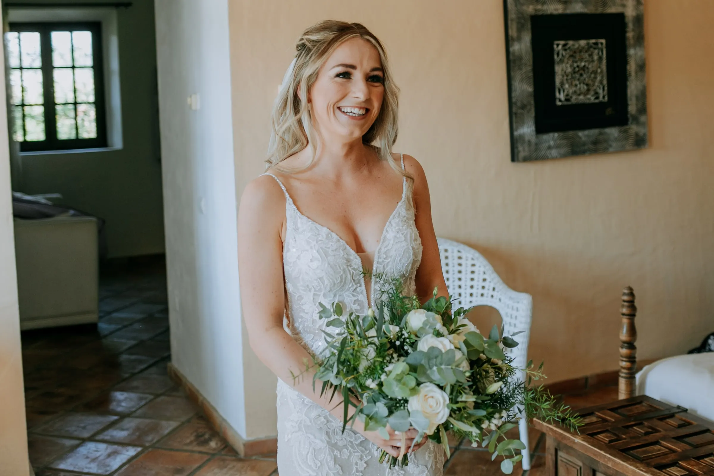 La novia, con vestido de encaje y ramo verde y blanco, sonríe en la habitación, Hacienda San José, Mijas