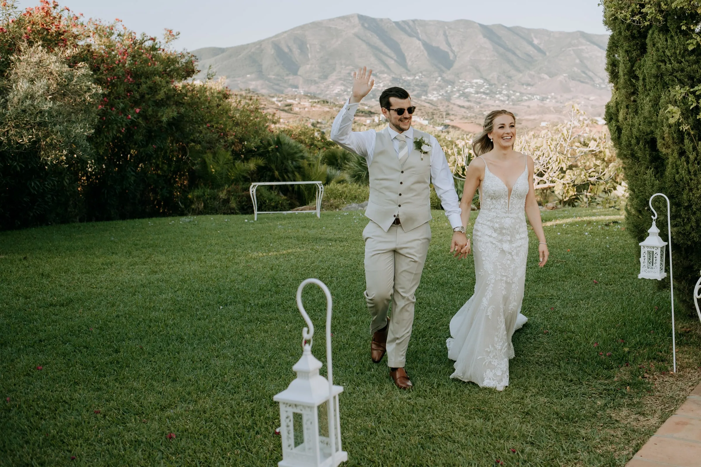 El novio saluda mientras camina con la novia por el césped entre farolillos blancos, Hacienda San José, Mijas
