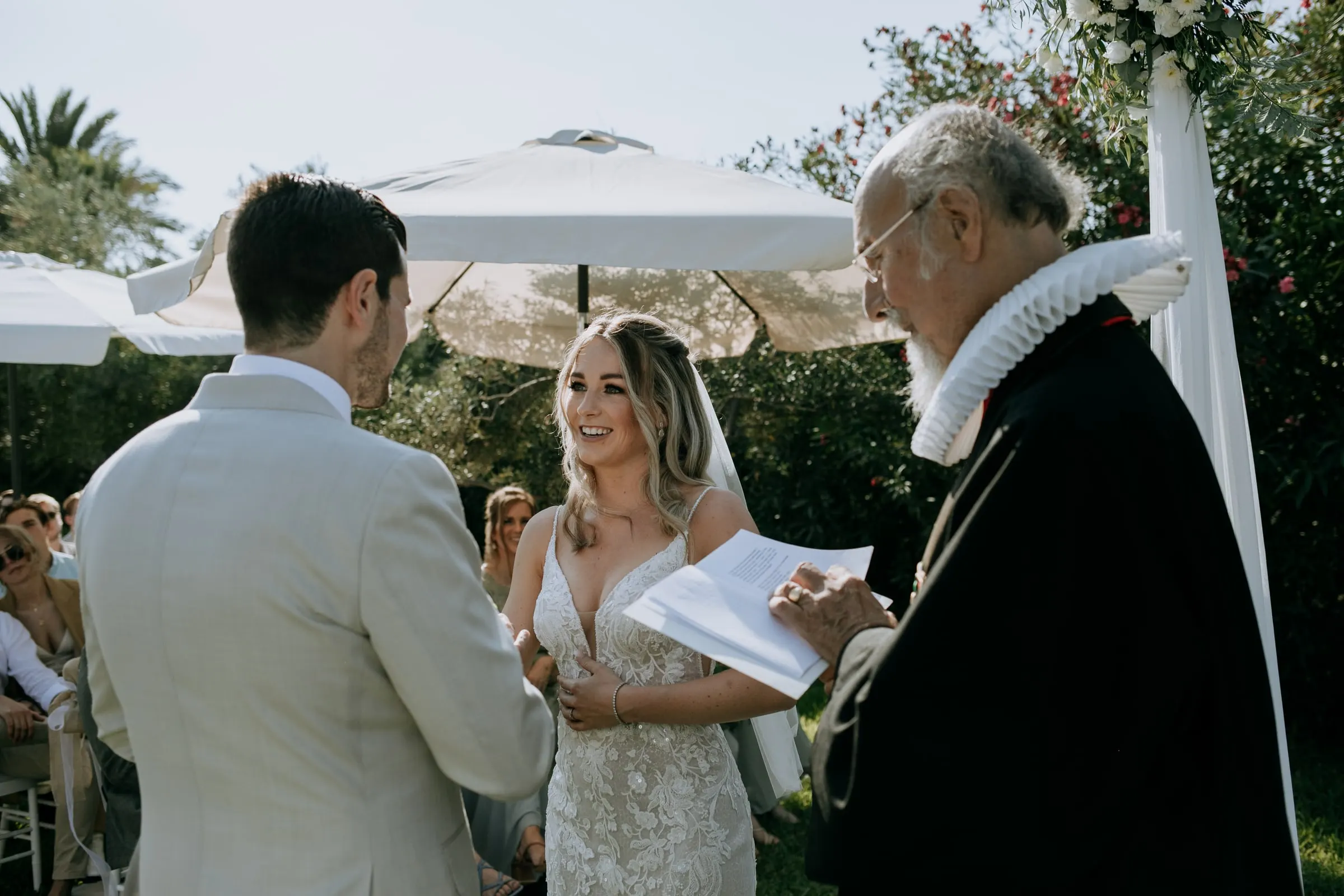 La novia sonríe mientras el oficiante, con gorguera, lee durante la ceremonia, Hacienda San José, Mijas