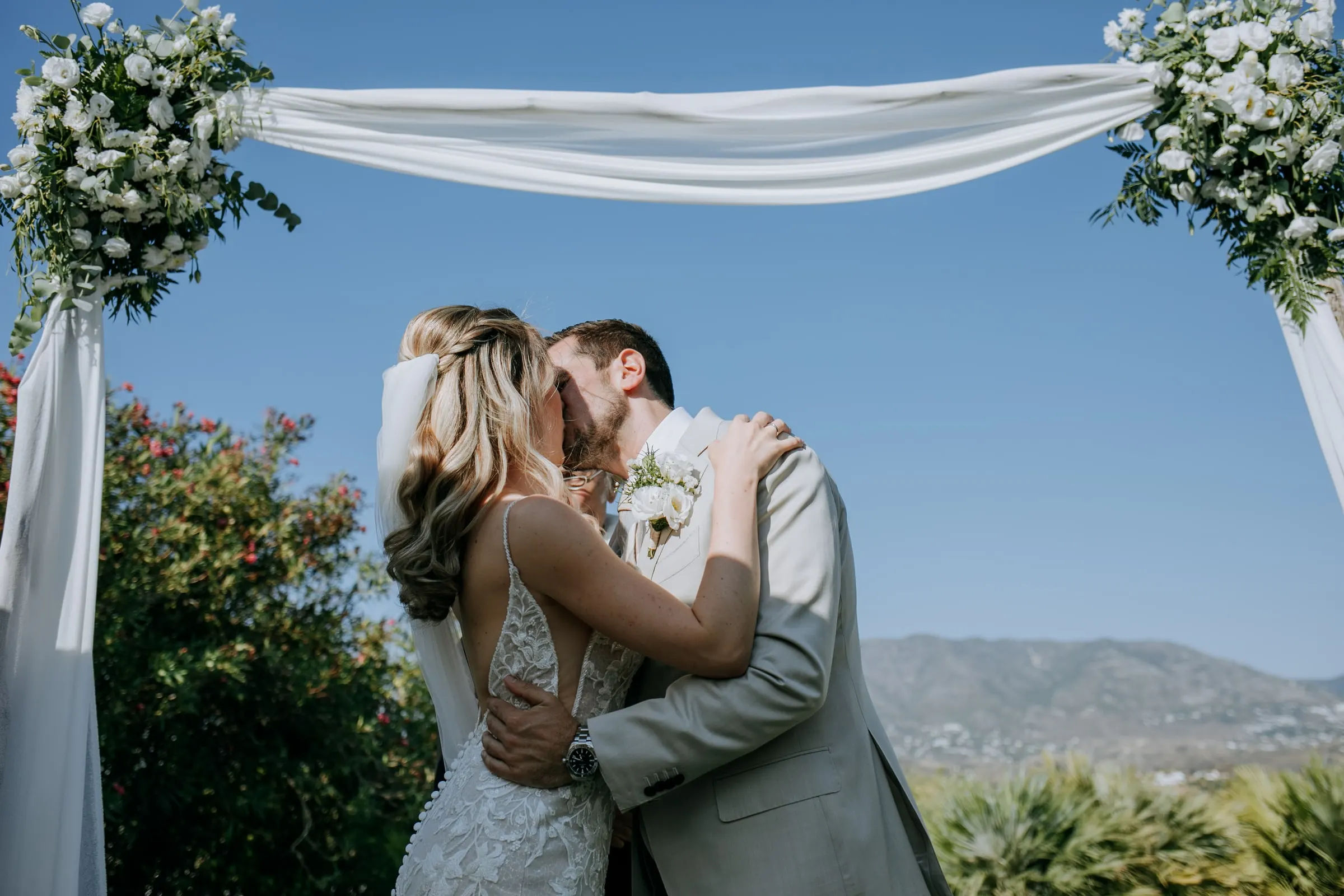 Los novios se besan bajo el arco de flores de la ceremonia con el cielo azul detrás, Hacienda San José, Mijas