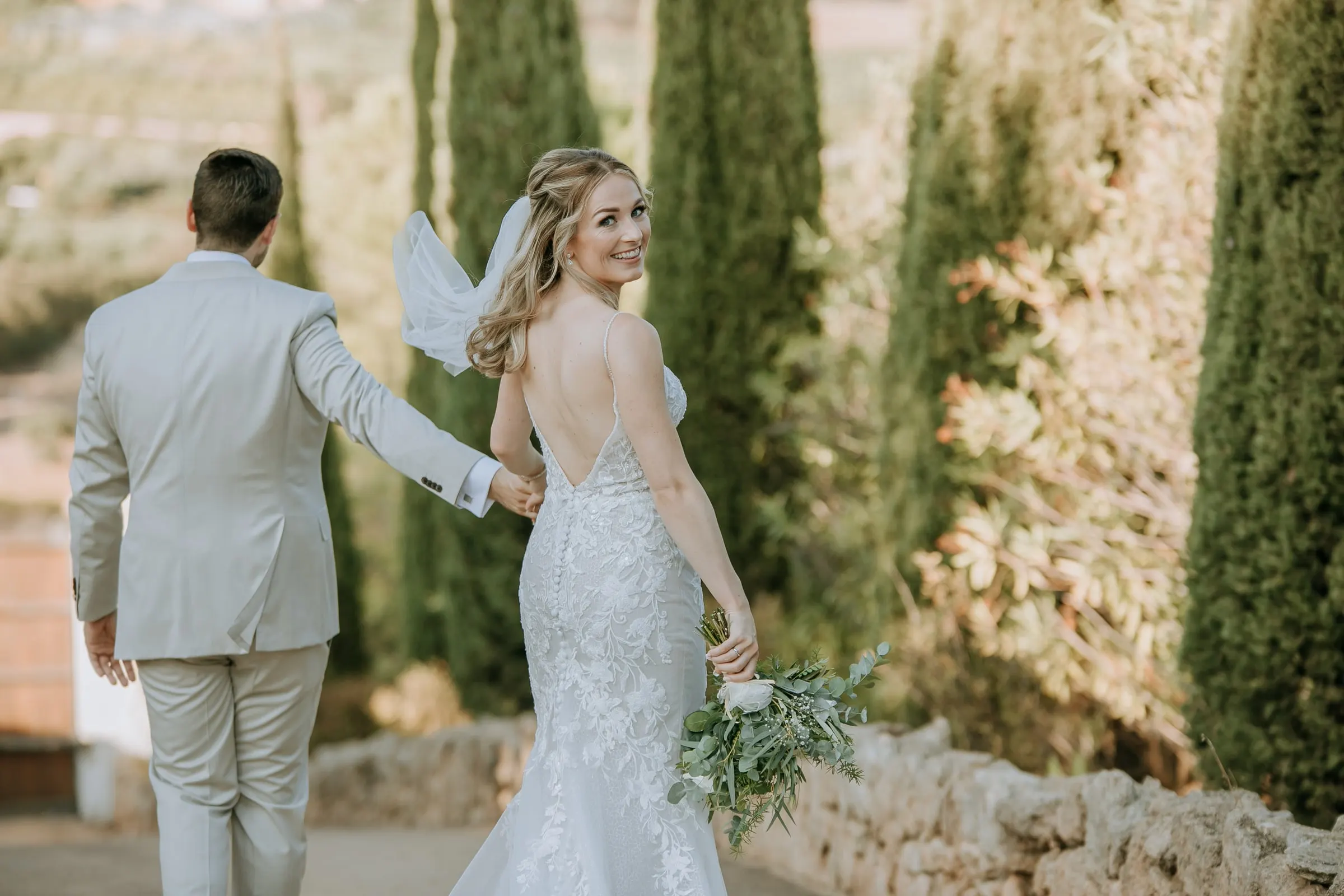 La novia mira atrás por encima del hombro mientras el novio la lleva entre cipreses, Hacienda San José, Mijas