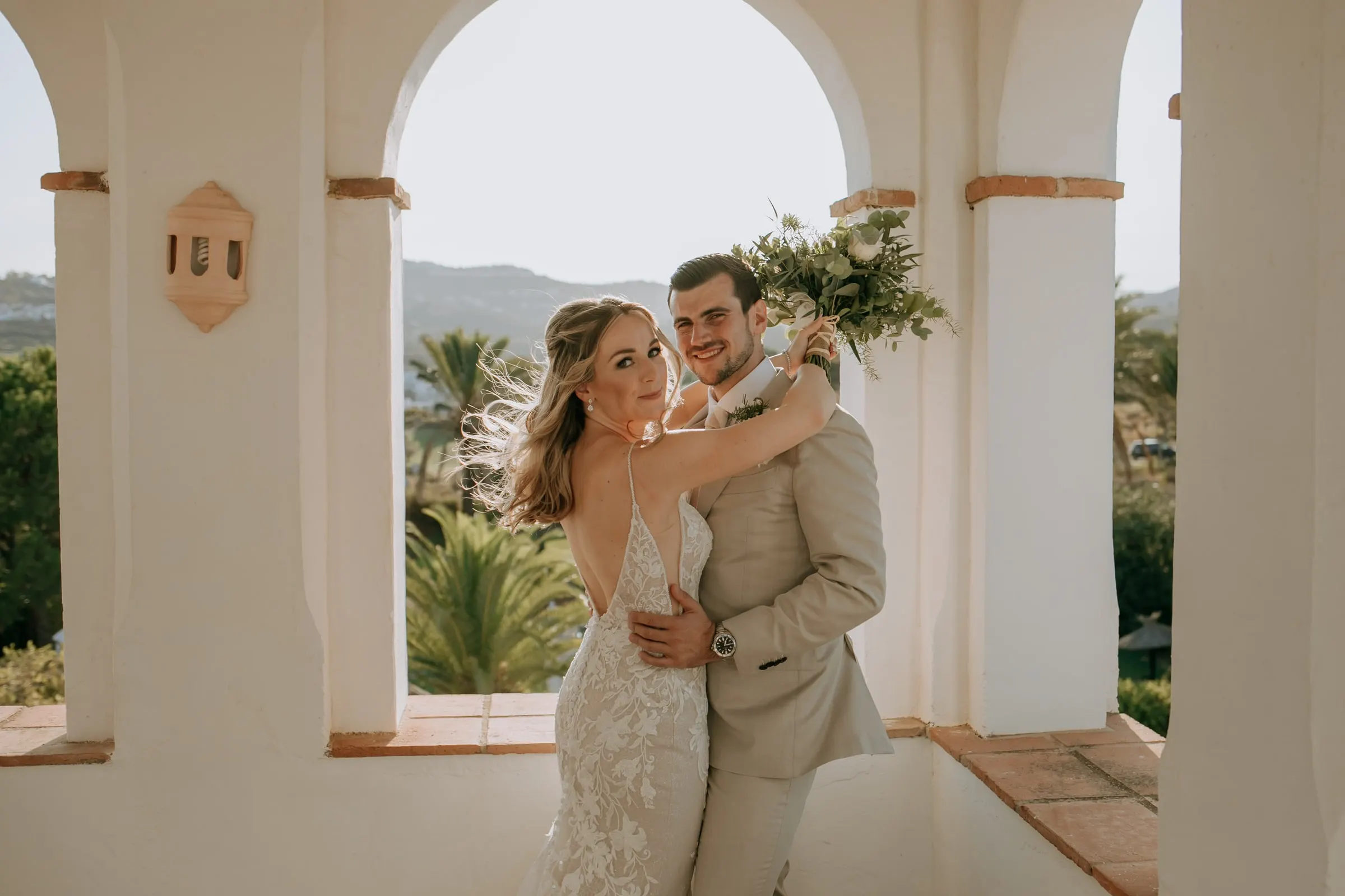 Los novios abrazados bajo los arcos blancos, con el ramo en alto y movido por la brisa, Hacienda San José, Mijas