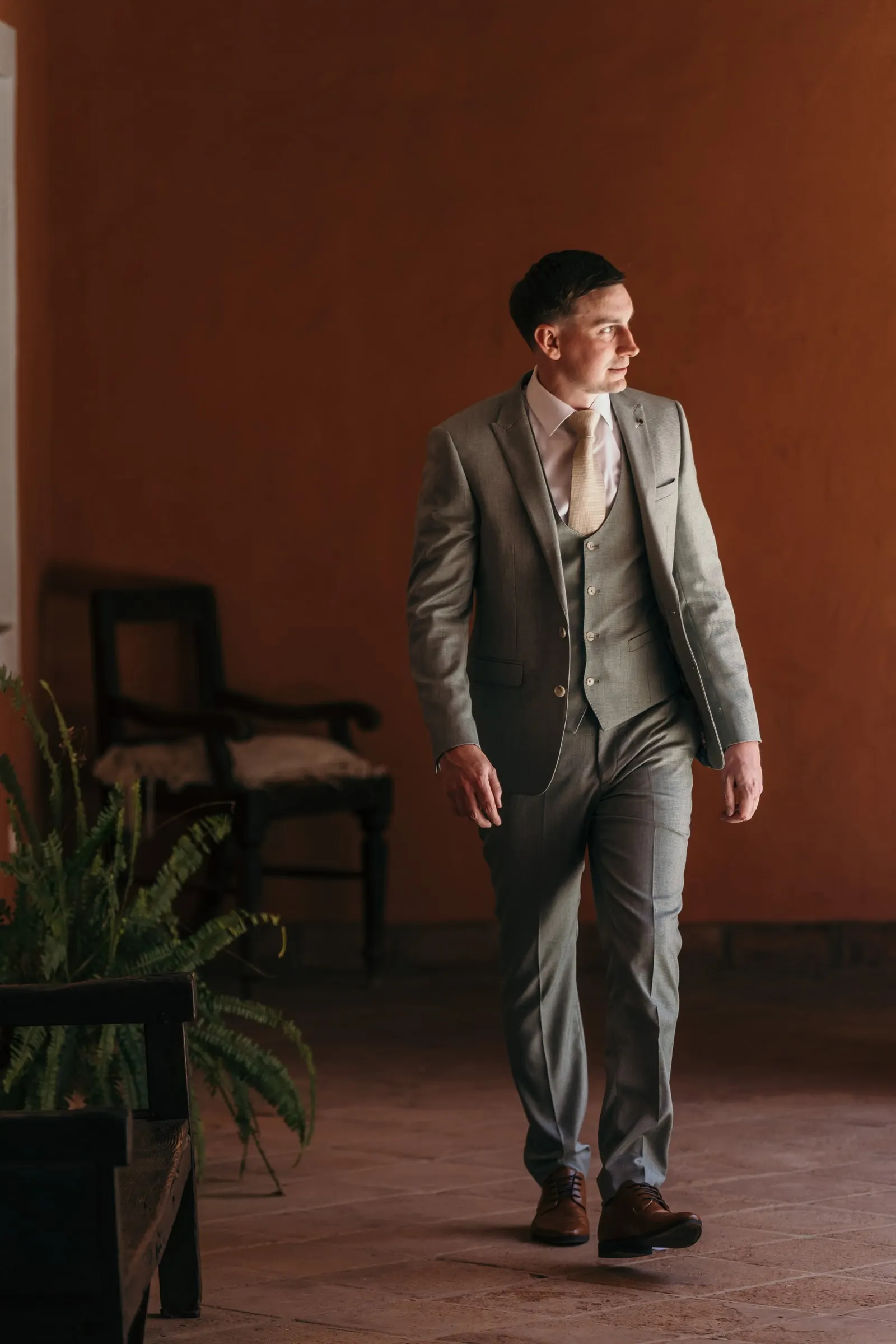 El novio, con traje gris de tres piezas, cruza una sala de suelo de barro