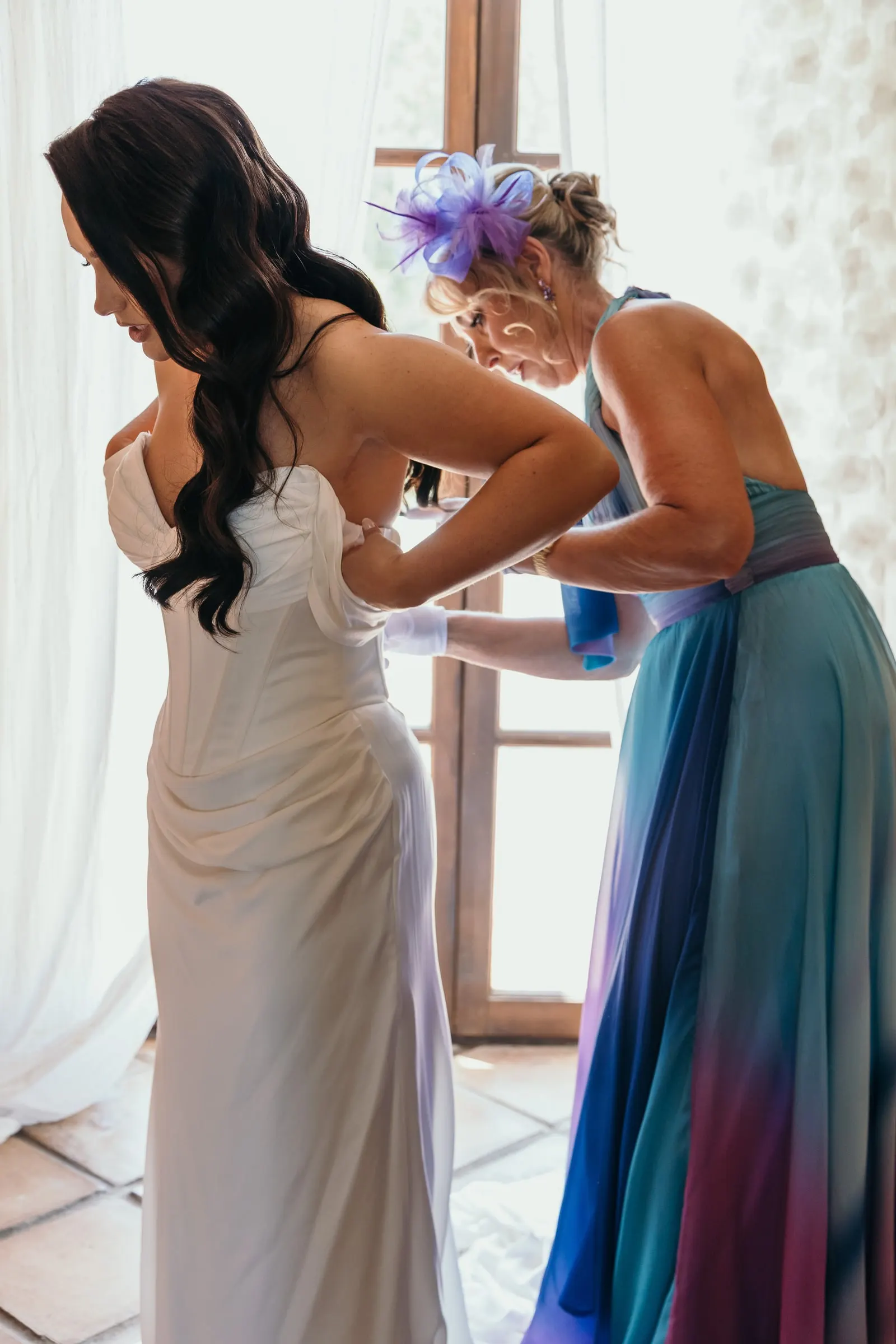 Una invitada abrocha la espalda del vestido de hombros caídos de la novia junto a la ventana