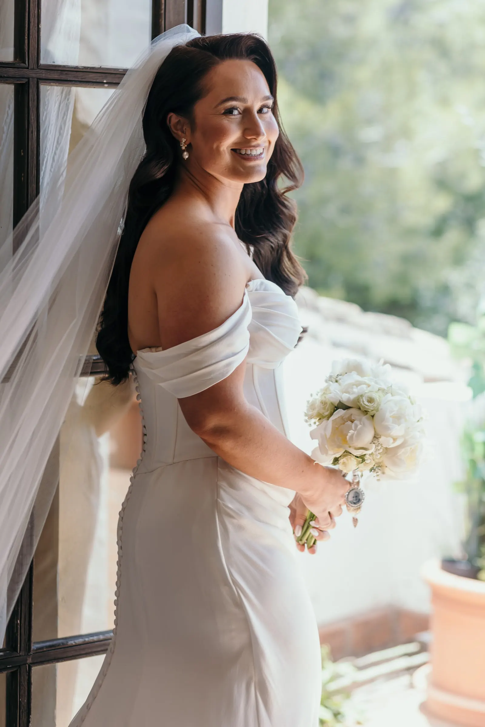 La novia, con su ramo de rosas blancas, mira por encima del hombro desde una puerta