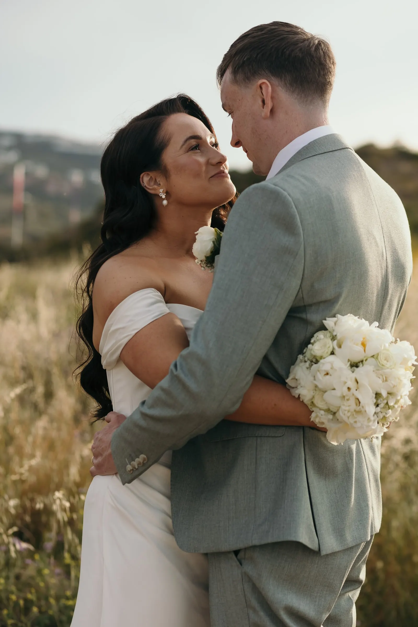 La novia abraza al novio con su ramo de rosas blancas entre la hierba alta