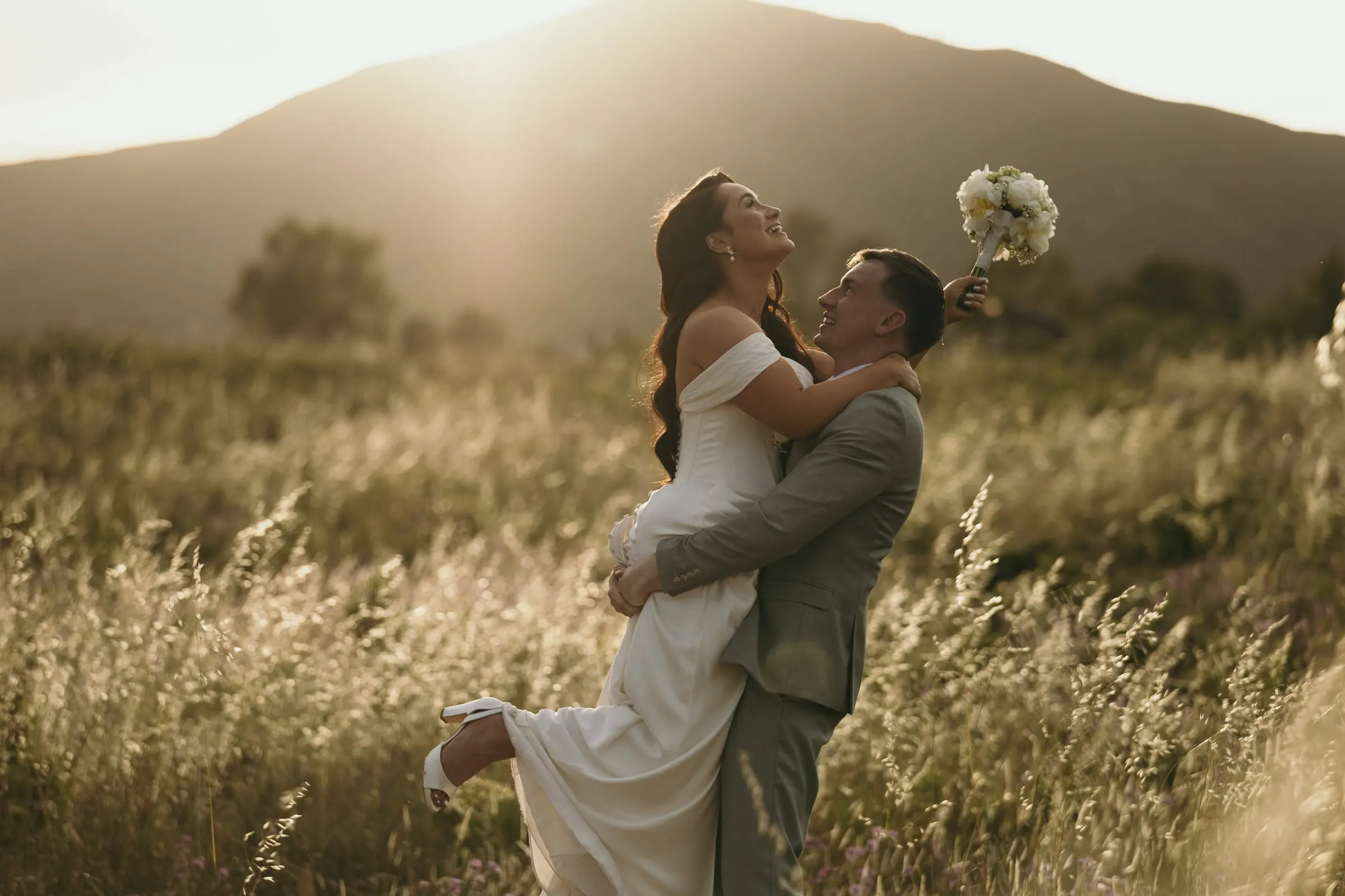 El novio levanta a la novia en un prado al atardecer, con la Sierra de Mijas detrás