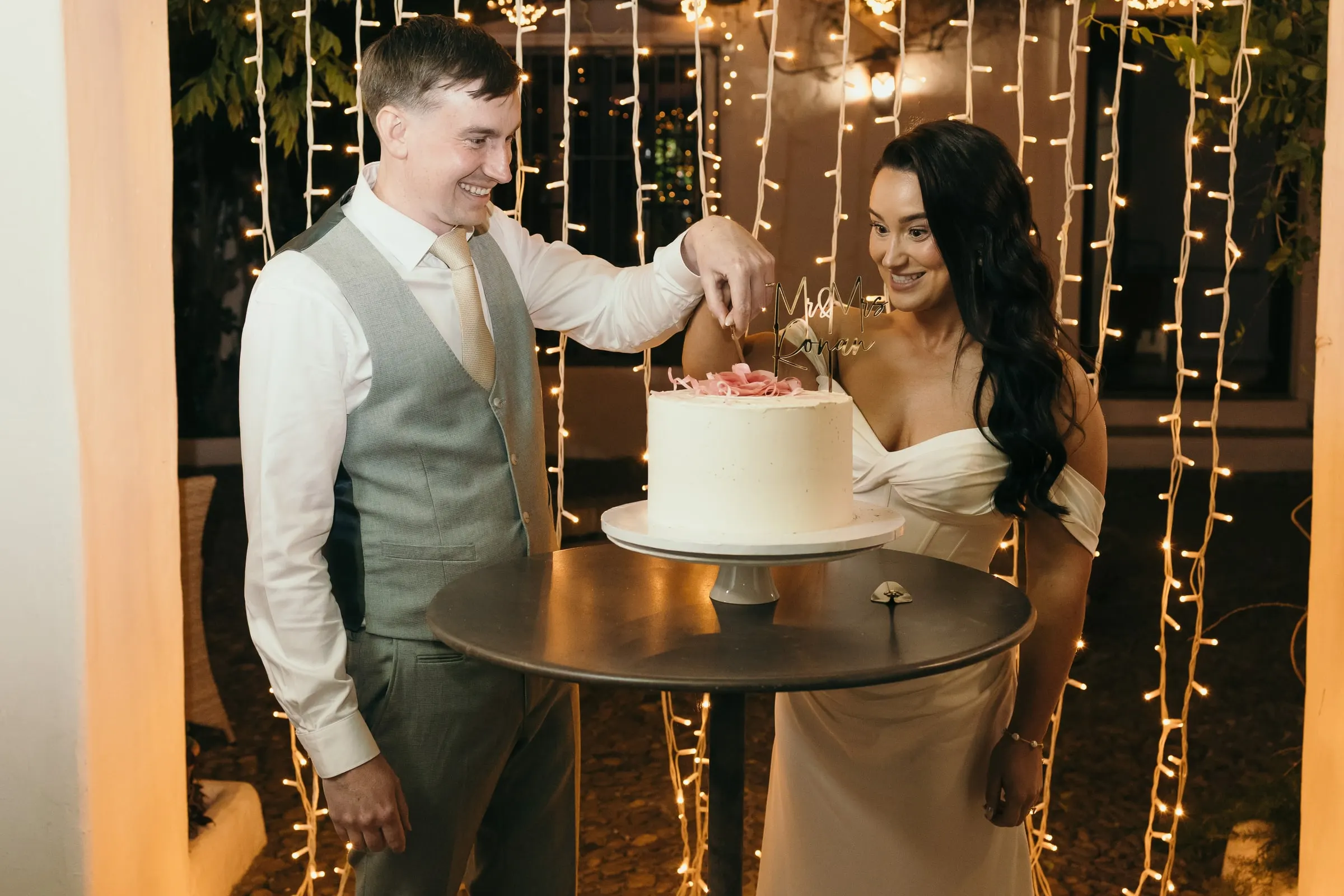 Los novios cortan una tarta blanca ante una cortina de lucecitas