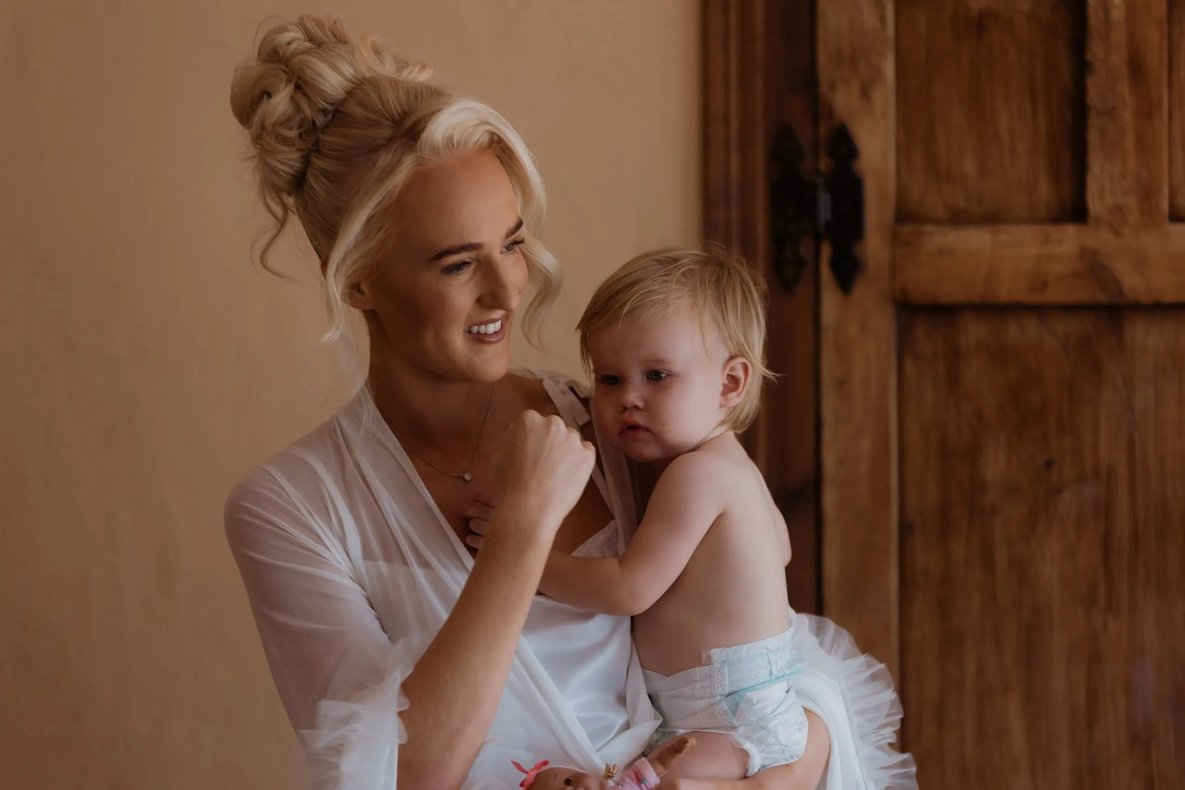Bride cuddling her baby by a wooden door on the wedding morning in Mijas