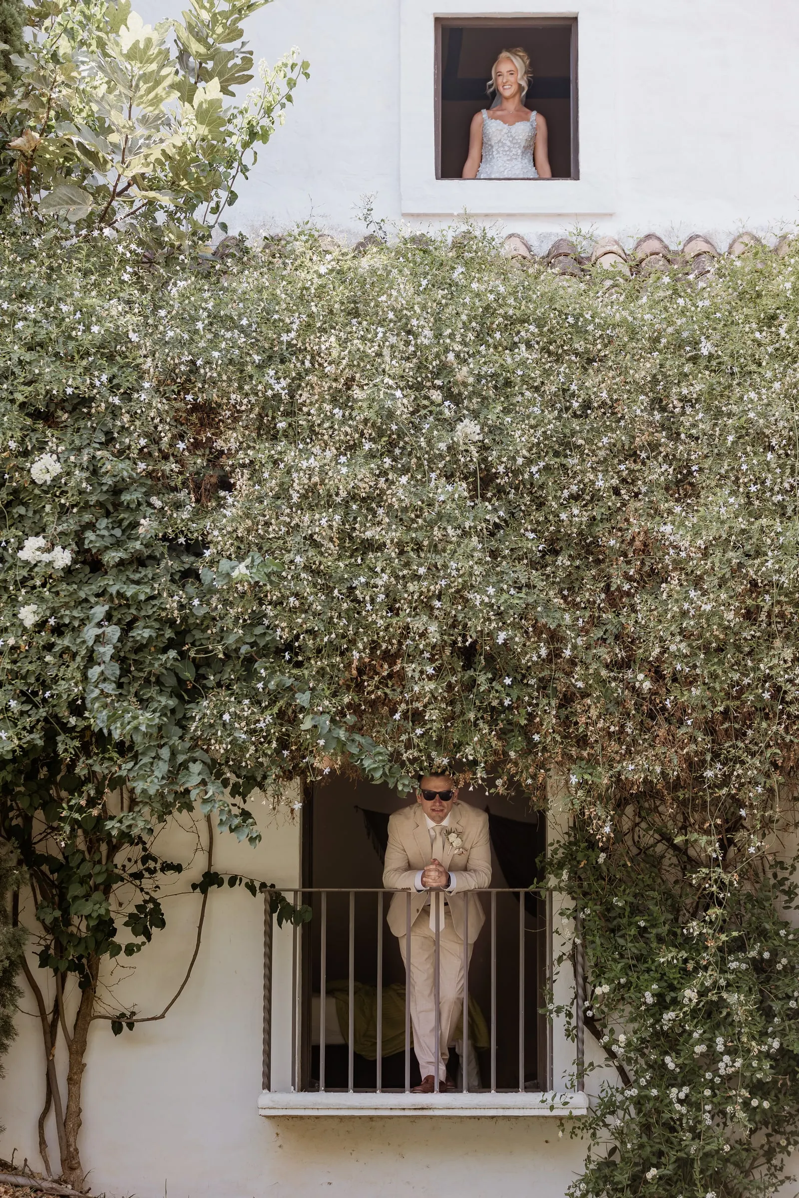 Bride at an upper window and a guest at the balcony below, on a white wall covered in climbing plants