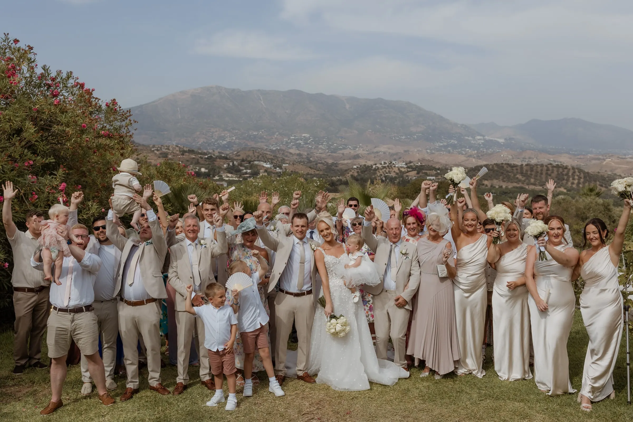 Big group photo of the wedding party cheering with the Mijas mountains behind