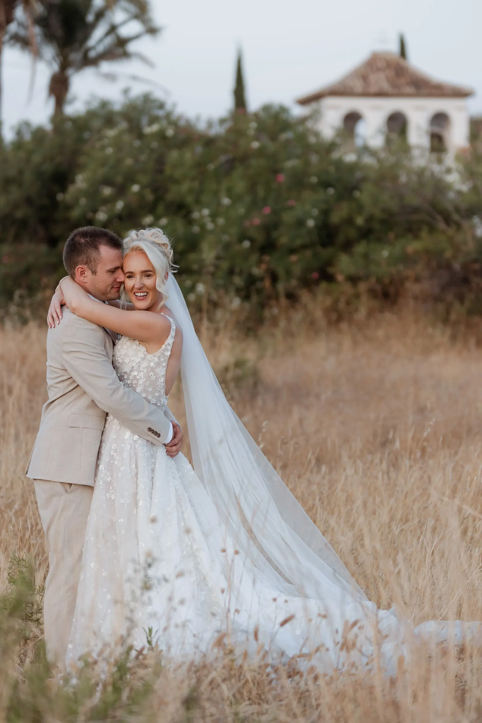 Los novios abrazados entre la hierba dorada con la torre blanca de Hacienda San José detrás