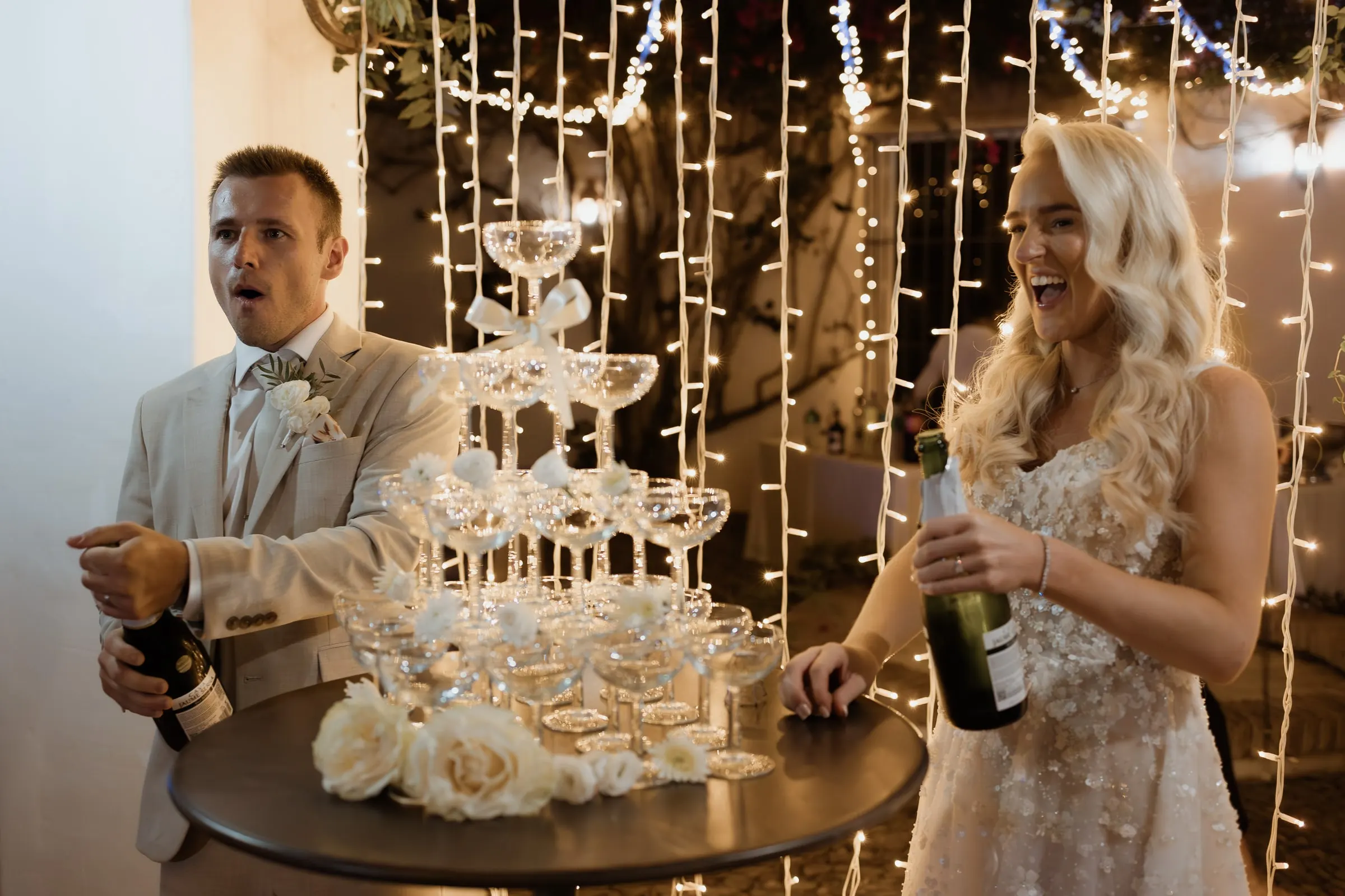 Bride and groom opening bottles beside a champagne tower under a curtain of lights