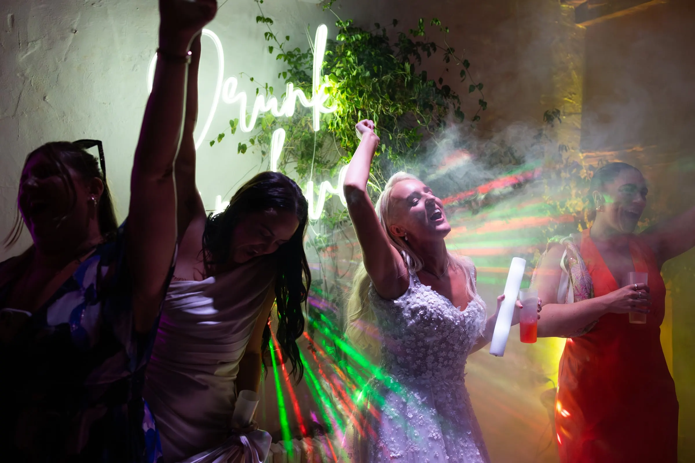 Bride and her friends dancing in the smoke beside a neon sign at Hacienda San José